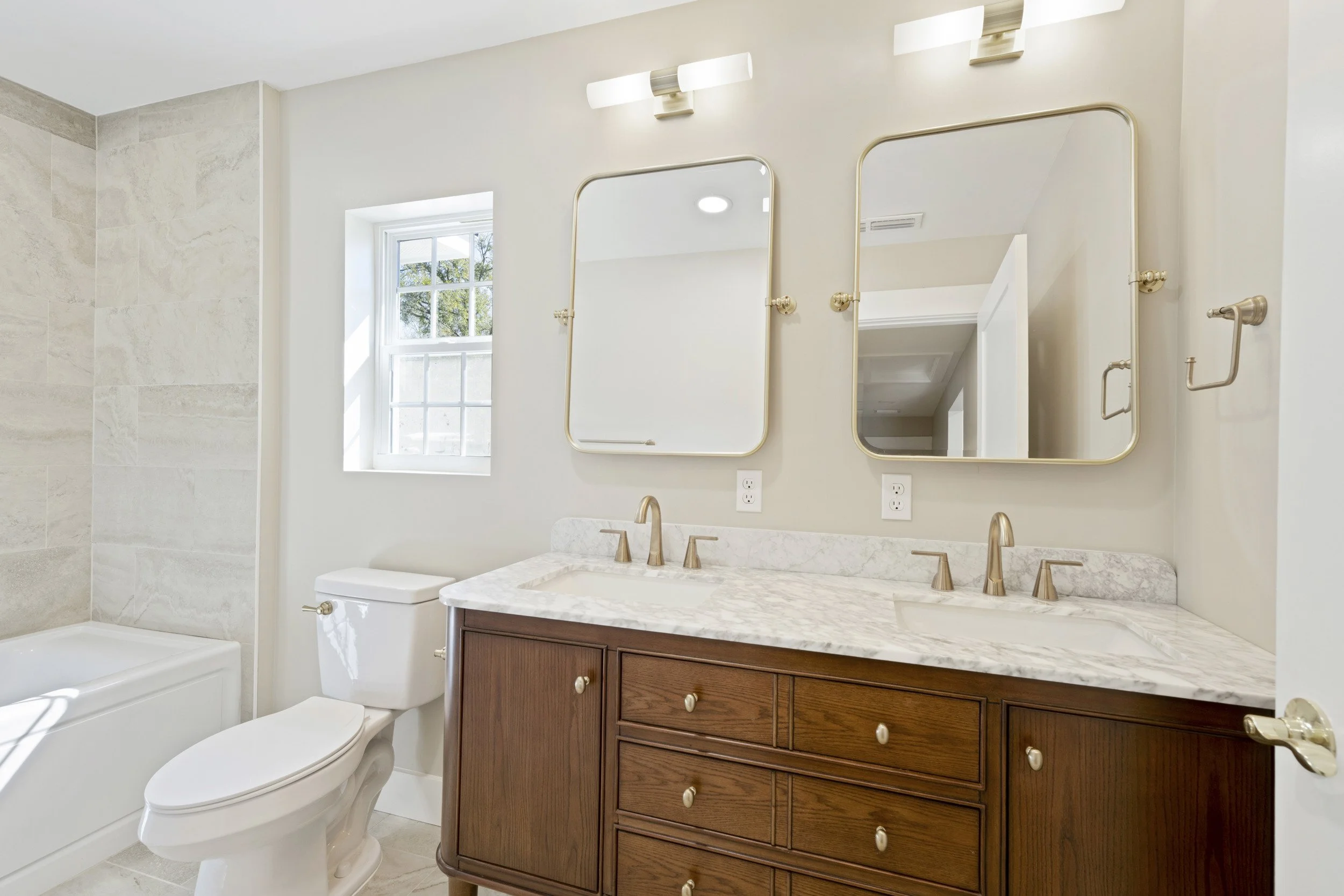 Bathroom with double sinks and marble countertop, two mirrors, light fixtures, toilet, bathtub, window, light-colored walls, and wooden vanity.