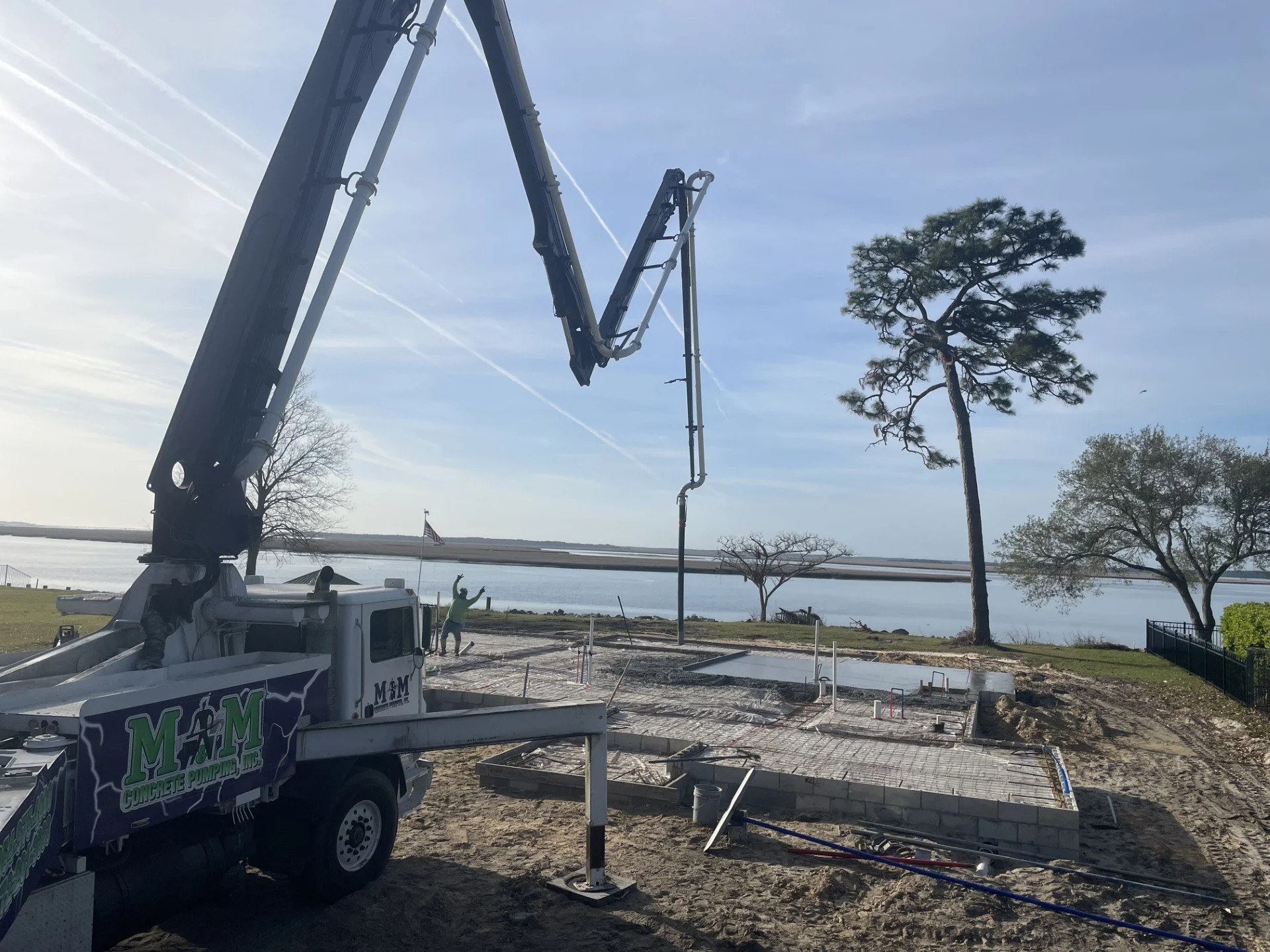 Construction site near water with a crane truck pouring concrete; trees and an American flag in the background.