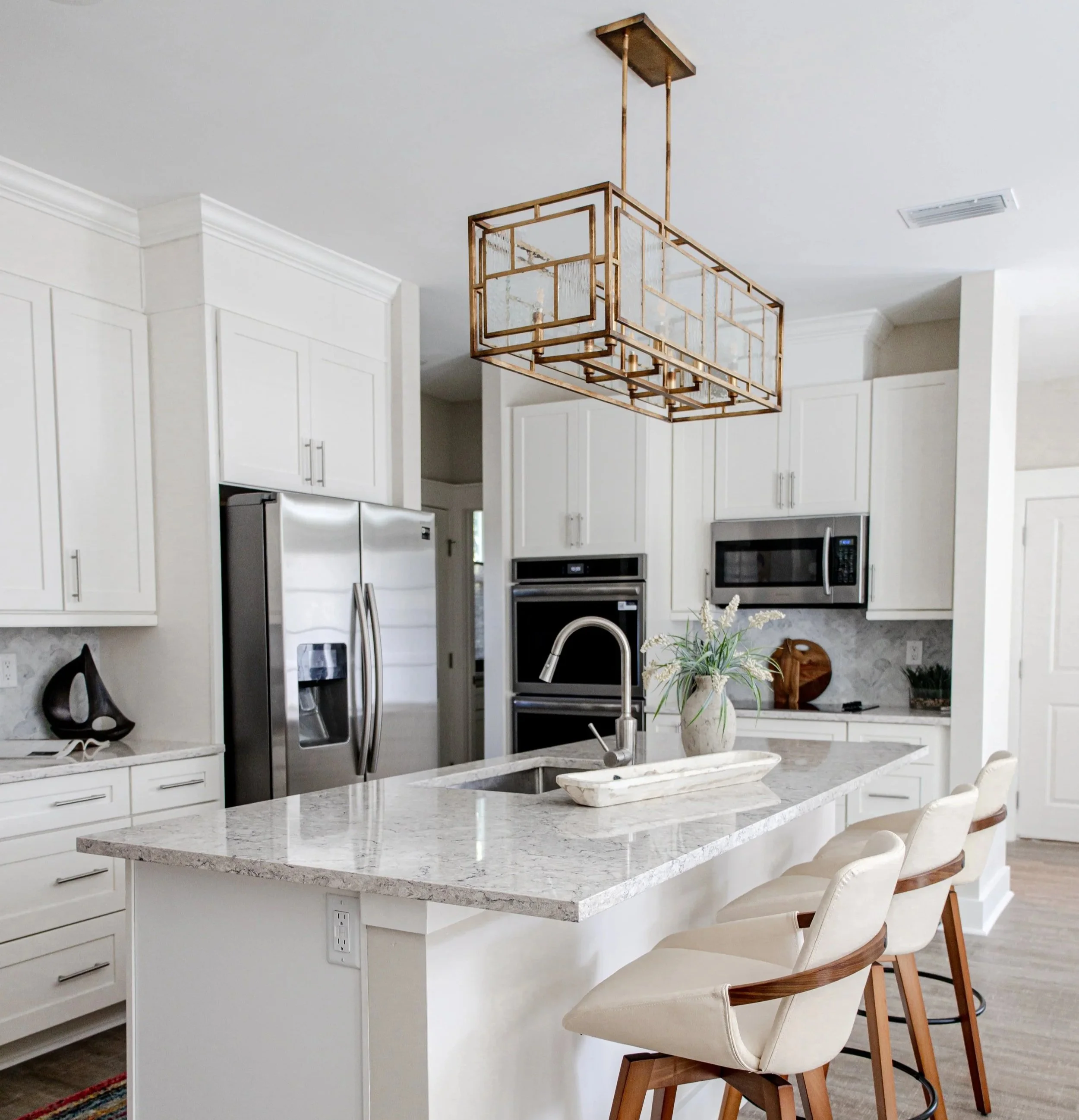 Modern kitchen with white cabinets, stainless steel refrigerator, marble island with sink, decorative gold chandelier, and beige bar stools.