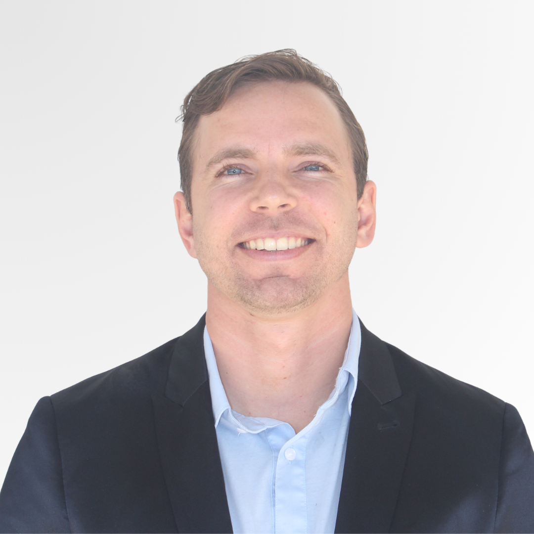 A man with short brown hair, blue eyes, and light skin wearing a light blue collared shirt and a black blazer, smiling against a plain white background.