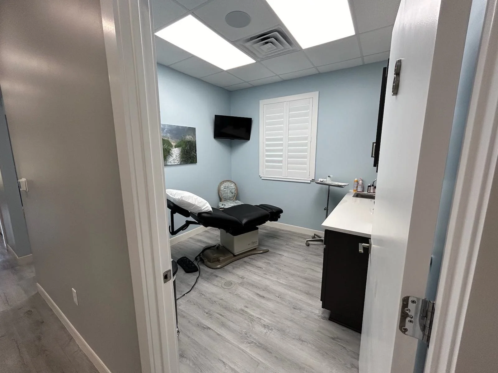 A medical examination room with a black examination table, a small chair, a mounted TV, a window with shutters, a cabinet, and various medical supplies.