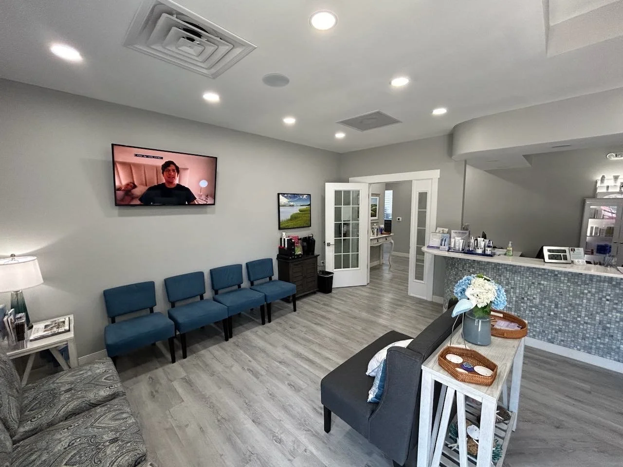 A waiting room with a row of five blue chairs, a small table with a lamp and magazines, a floral arrangement on a side table, a reception desk with various items, a wall-mounted television showing a man, and a doorway with glass panels leading to another room.
