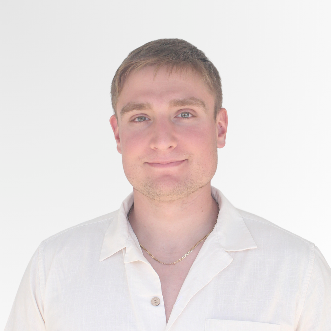 A young man with short light brown hair, blue eyes, and light skin, smiling slightly, wearing a white button-up shirt and a gold chain necklace, against a plain white background.