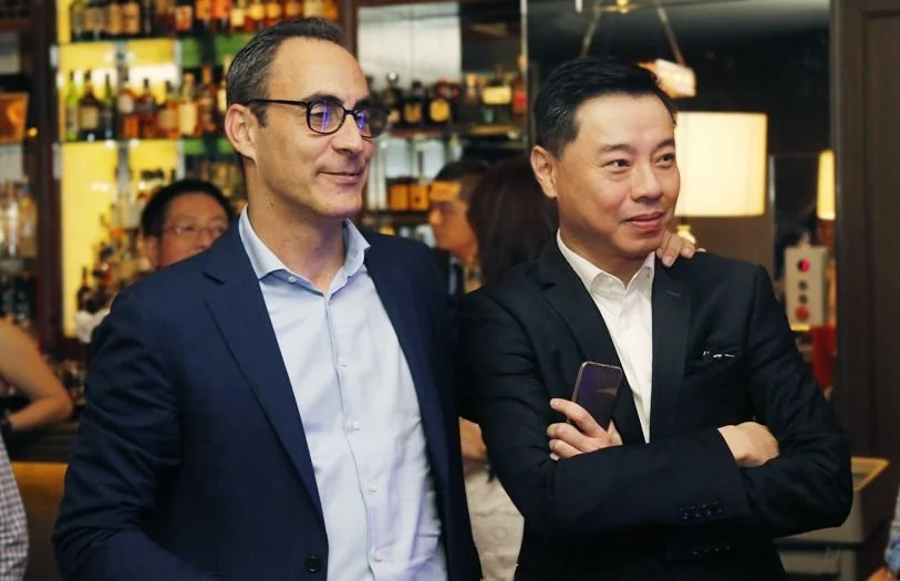 Two men in suits standing in a bar or restaurant, with drinks and bottles on shelves behind them, and other people in the background.