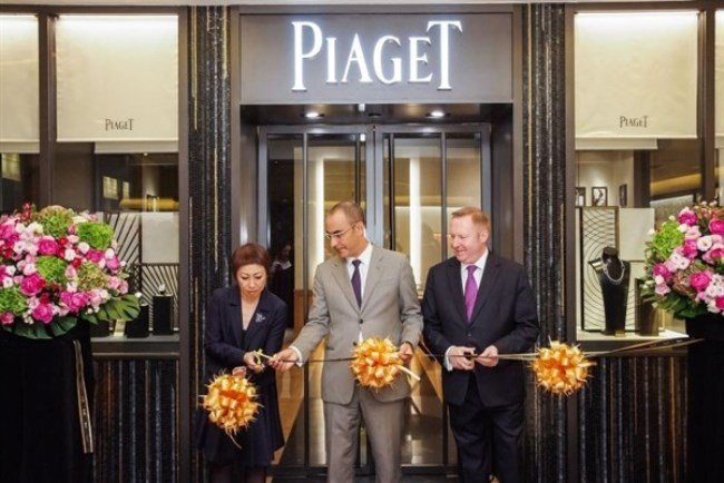 Three people in formal attire participating in a ribbon-cutting ceremony outside a Piaget store, with large flower arrangements on either side.