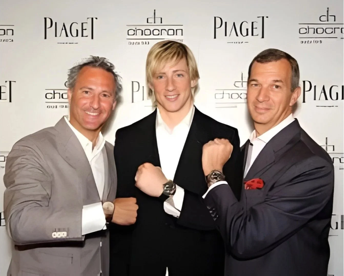 Three men in suits posing with their fists raised in front of a backdrop with logos for Piaget and Chopard, smiling at the camera.