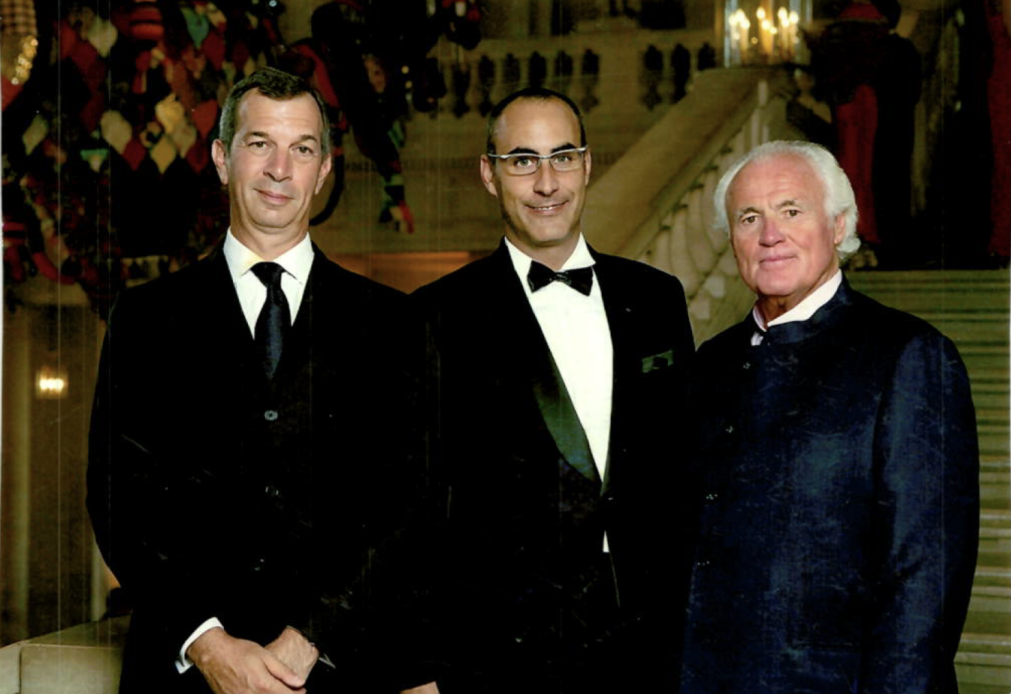 Three men in tuxedos standing inside a grand hall with a staircase in the background.