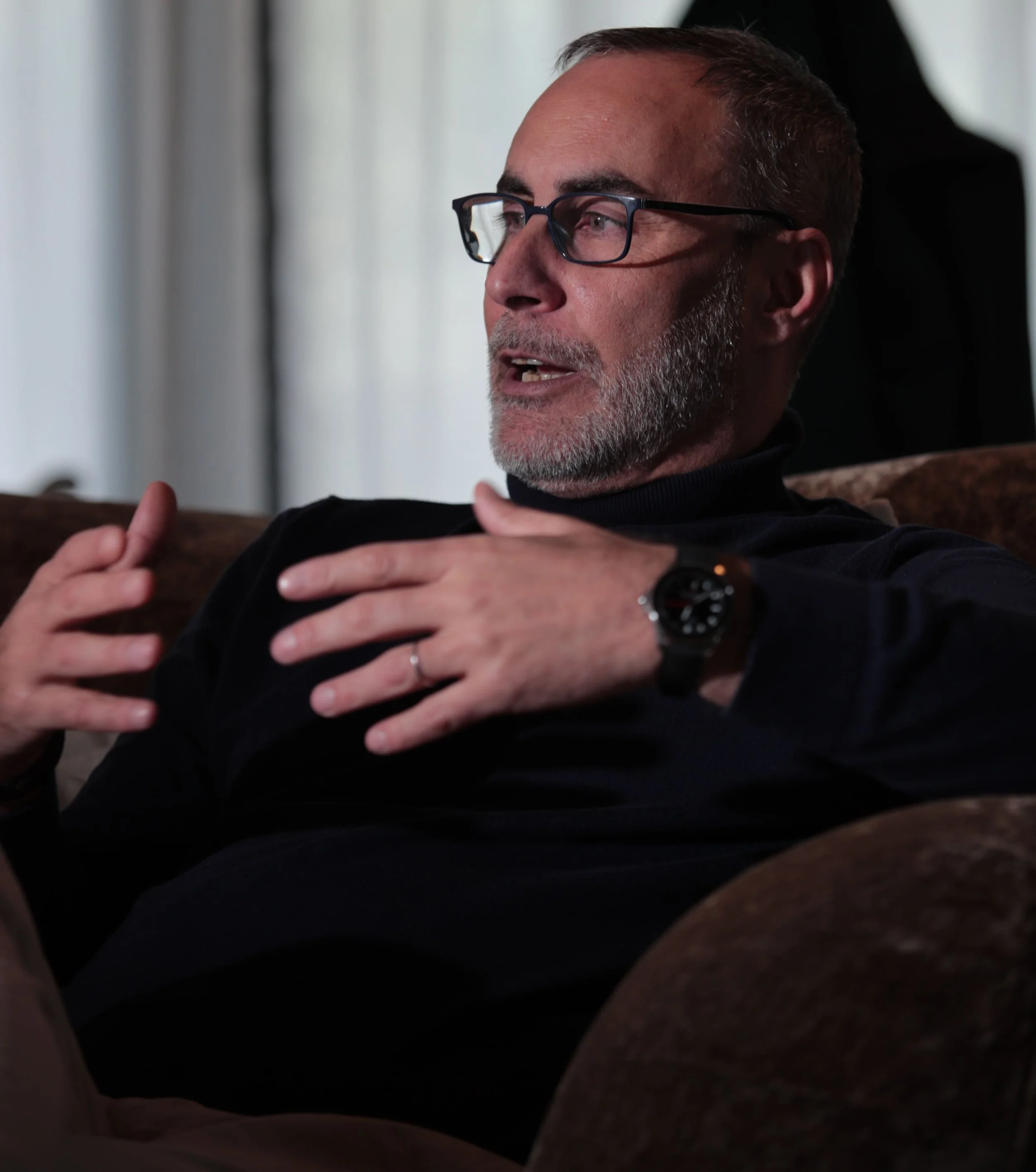 A middle-aged man with glasses, a beard, and a black turtleneck sweater sitting on a couch, speaking and gesturing with his hands.