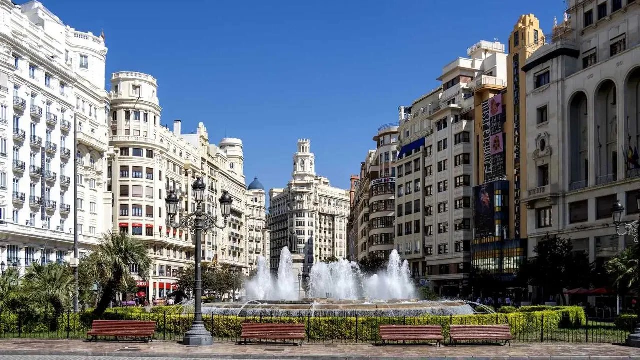 City square with a fountain, surrounded by tall historic buildings, benches, and trees, under a clear blue sky.