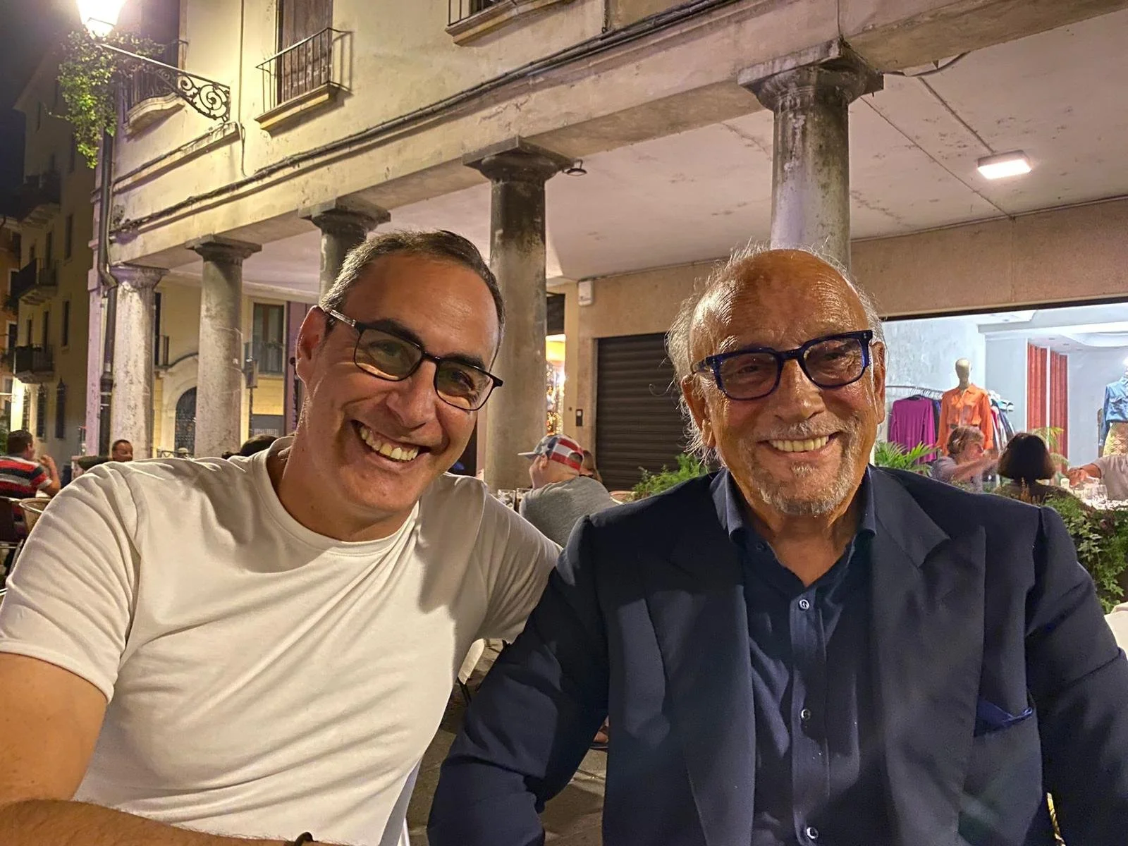Two men smiling and sitting together at an outdoor restaurant or cafe during the evening, with buildings and other people in the background.
