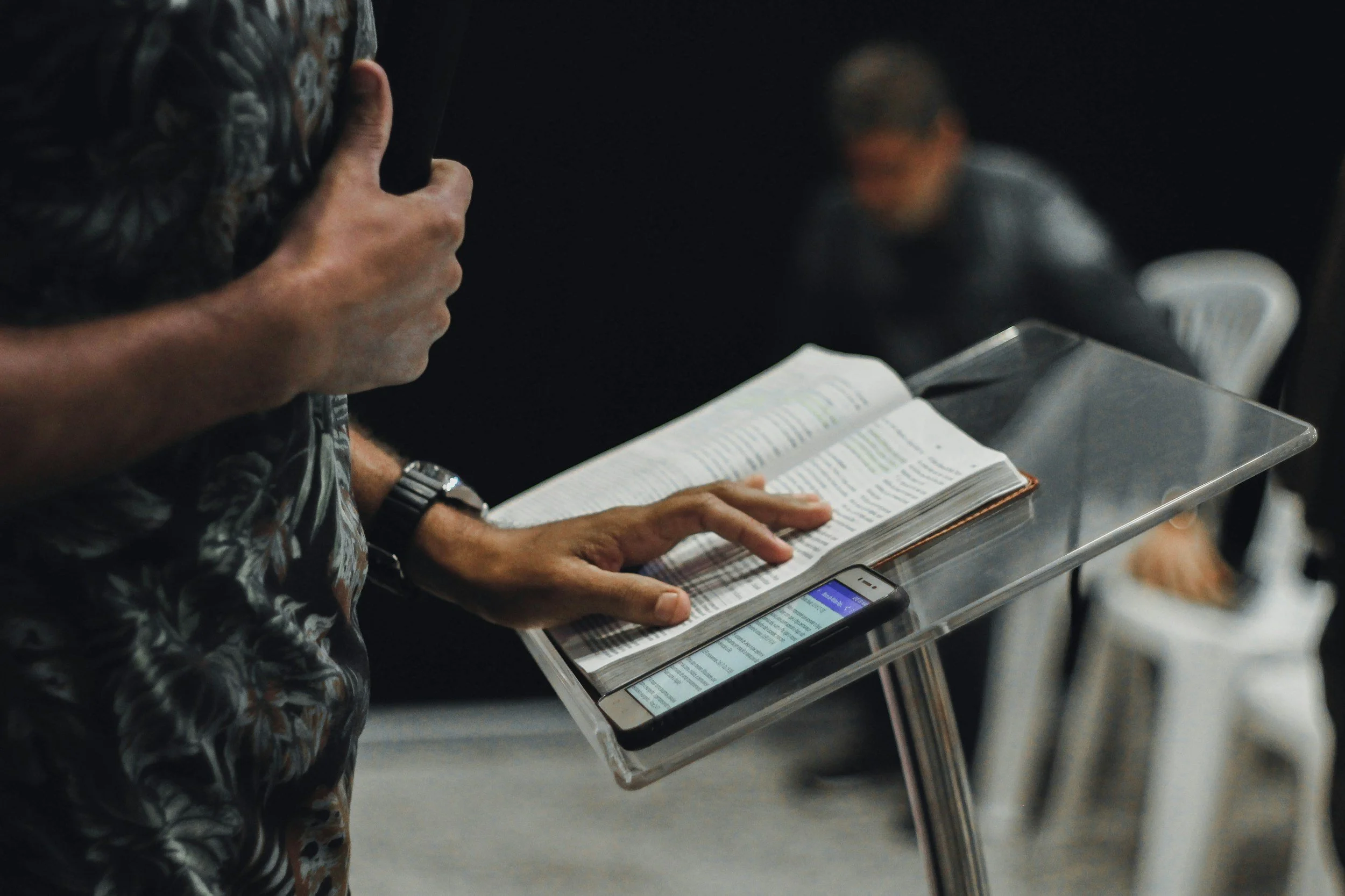 Person standing at a clear plastic lectern reading from a large book and using a smartphone, with another person blurred in the background.