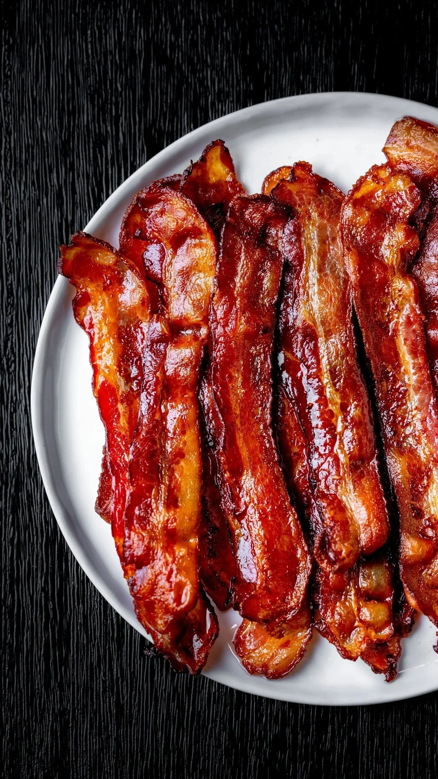 Men's breakfast. A white plate with several cooked strips of bacon on a black wooden surface.