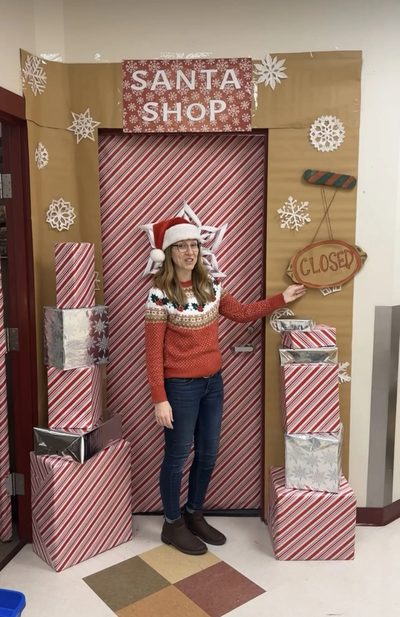 A woman dressed in a Christmas sweater and Santa hat standing in front of a festive holiday display with a 'Santa Shop' sign, snowflake decorations, and wrapped presents, while holding a 'Closed' sign.