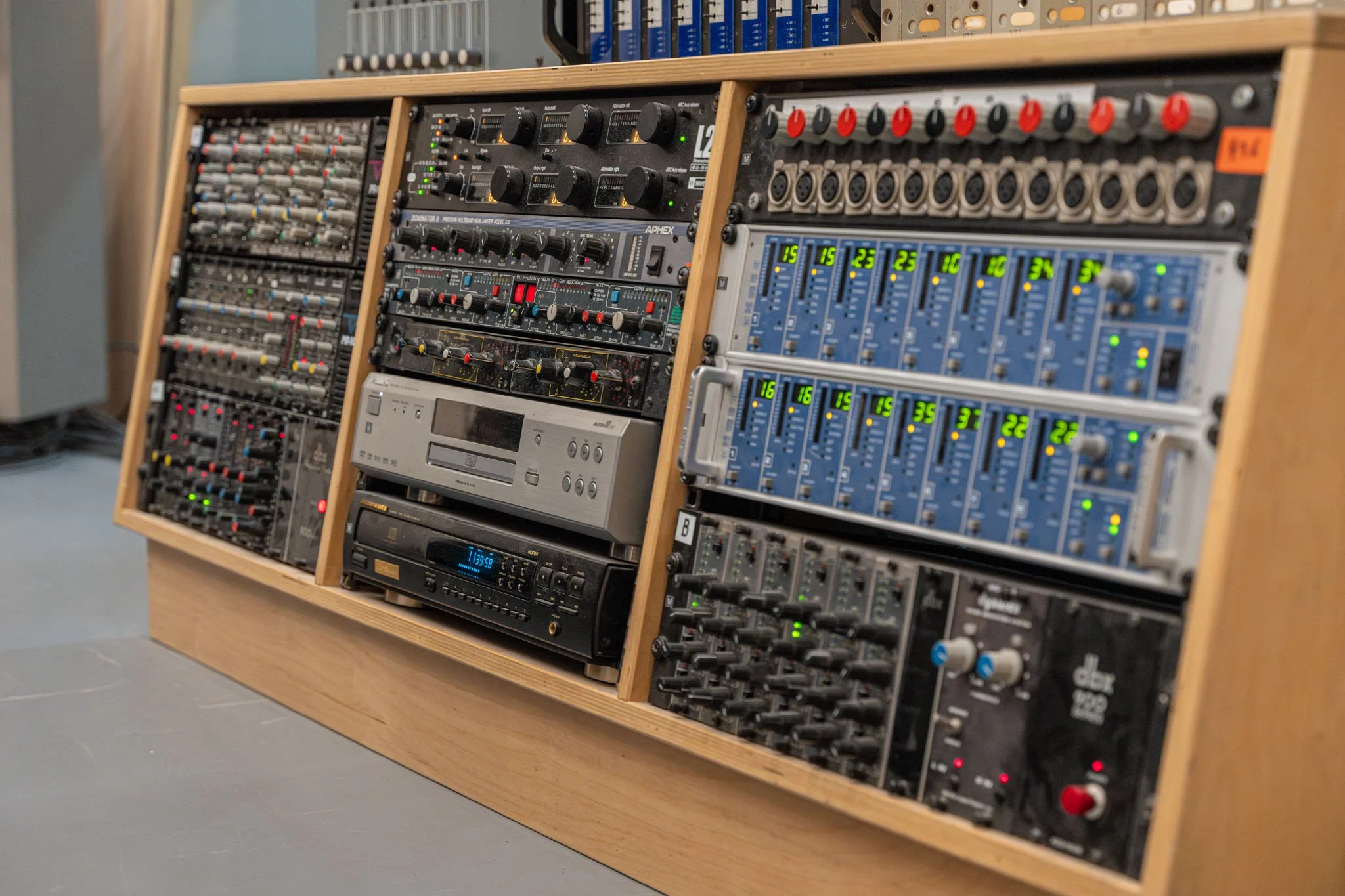 A wooden rack holding multiple audio equipment units including mixers, equalizers, and audio processors in a recording studio.
