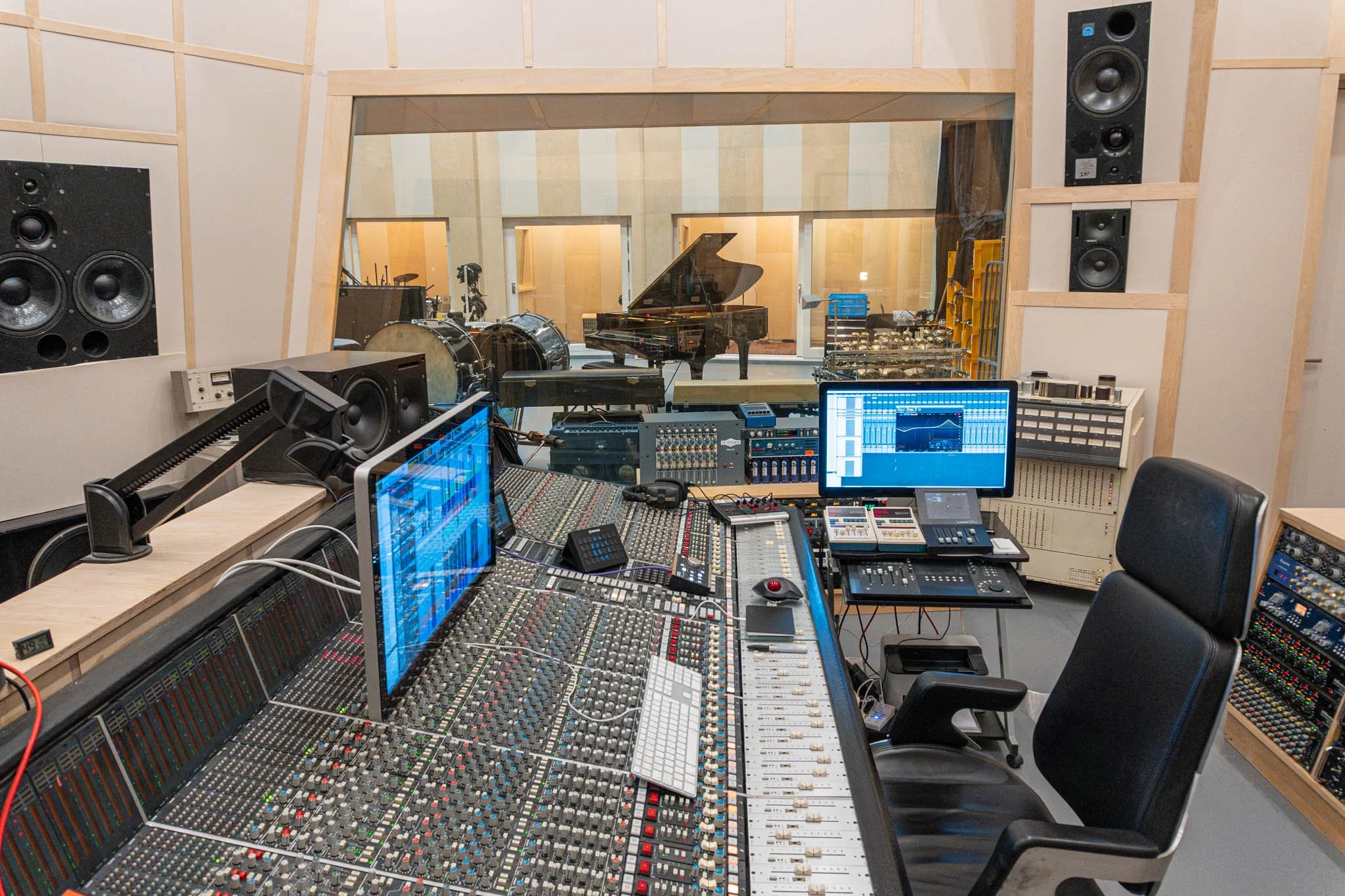 Inside a professional recording studio showing a mixing console, two computer screens, microphones, and musical instruments including drums and a grand piano, with soundproof walls and large studio speakers.