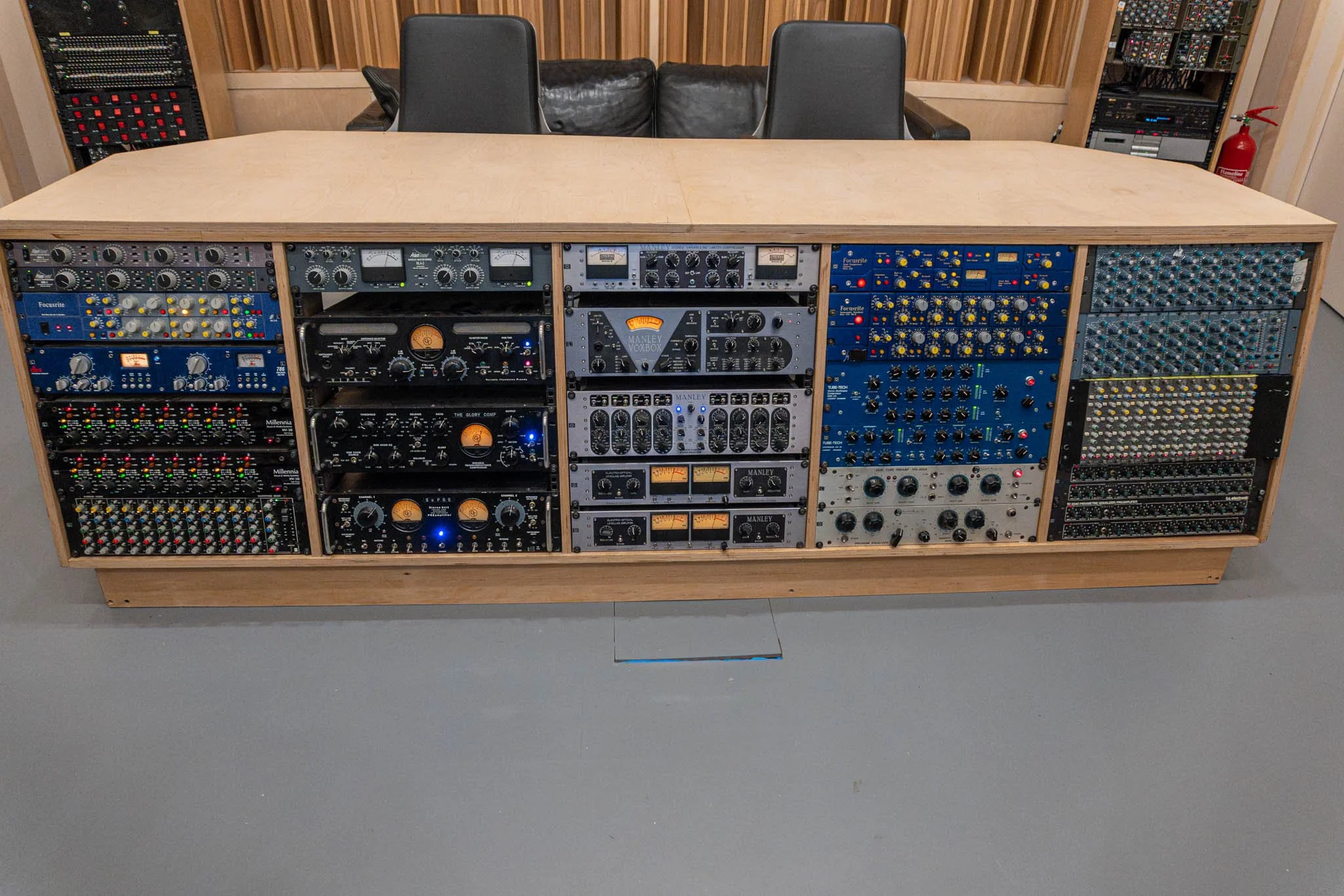 A large wooden mixing console with various audio equipment and hardware patches, situated in a recording studio with chairs and acoustic paneling in the background.