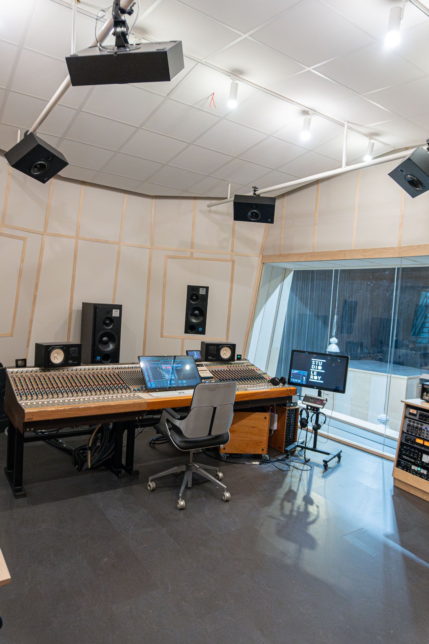 A professional recording studio with a large mixing console, six studio monitor speakers mounted on the ceiling for 7.2.4 Atmos, and a window looking into an adjacent room.
