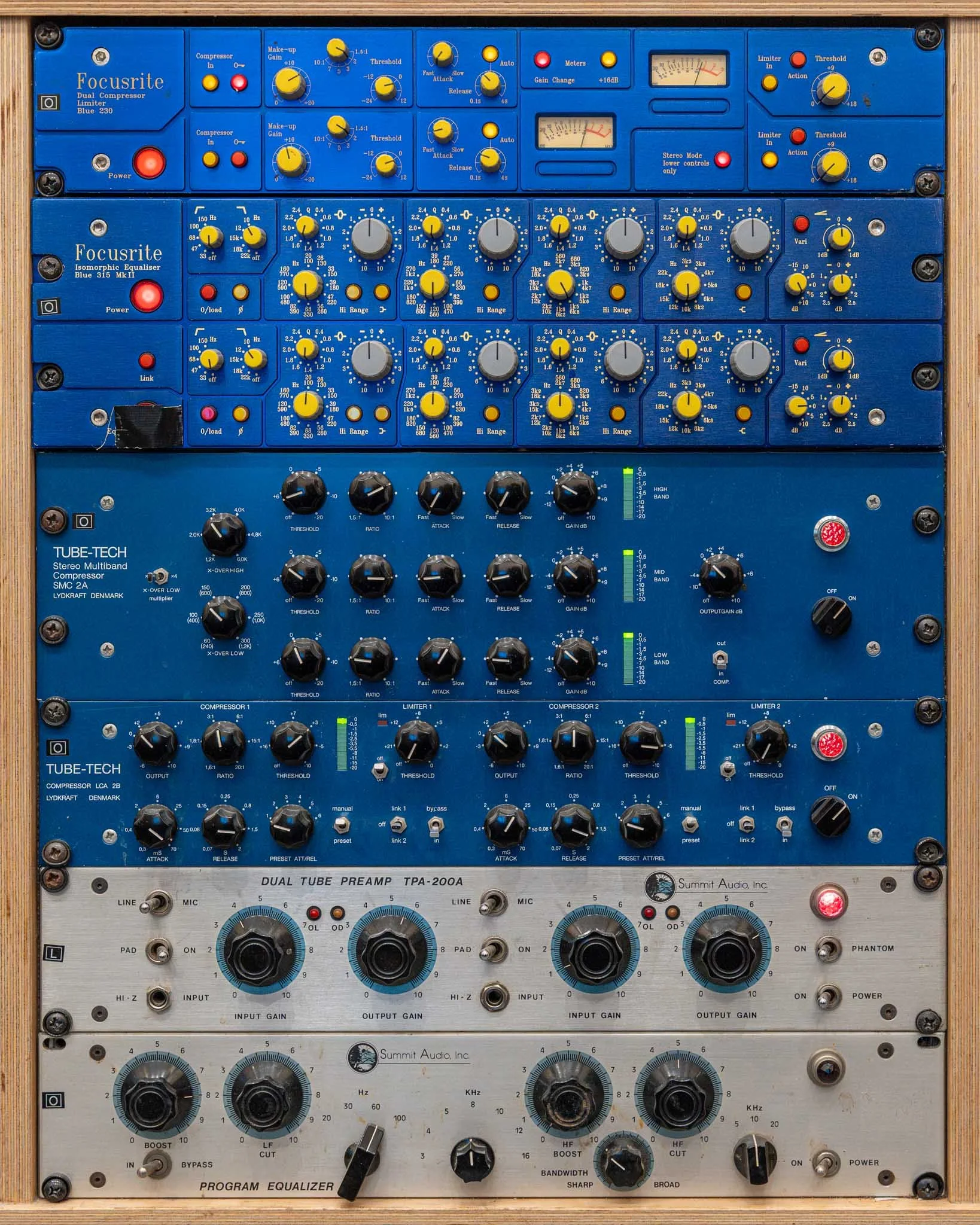 Audio equipment rack with multiple blue and silver units, including compressors, equalizers, preamps, and a program equalizer, with various knobs, switches, and meters.