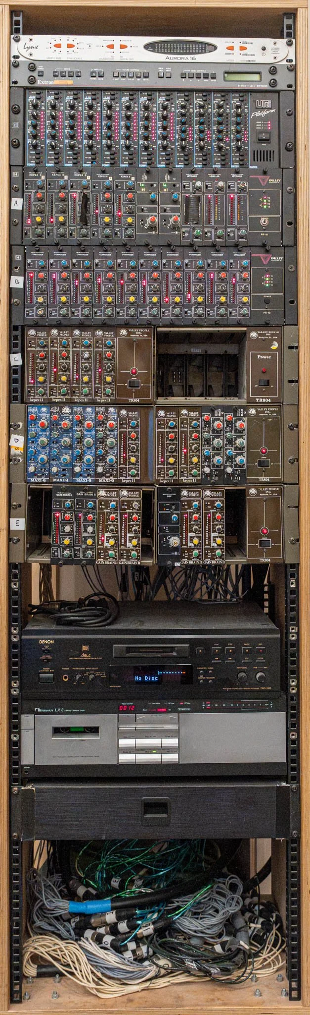 A tall audio equipment rack containing various audio mixing consoles, amplifiers, and a CD player, with numerous cables and wires at the bottom.