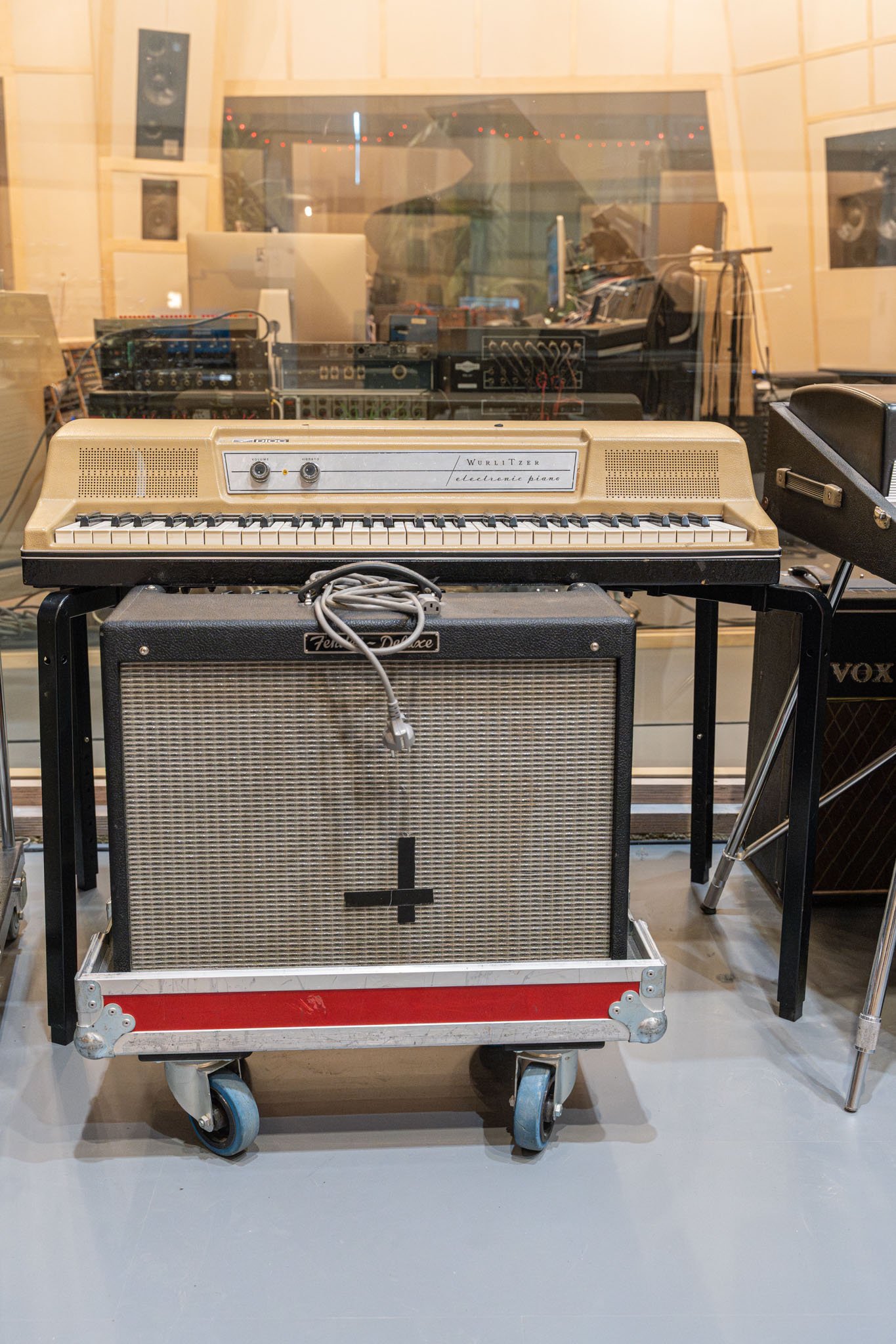 Vintage electronic keyboard on top of an amplifier, with a guitar amplifier to the right and studio equipment reflected in a glass window behind.