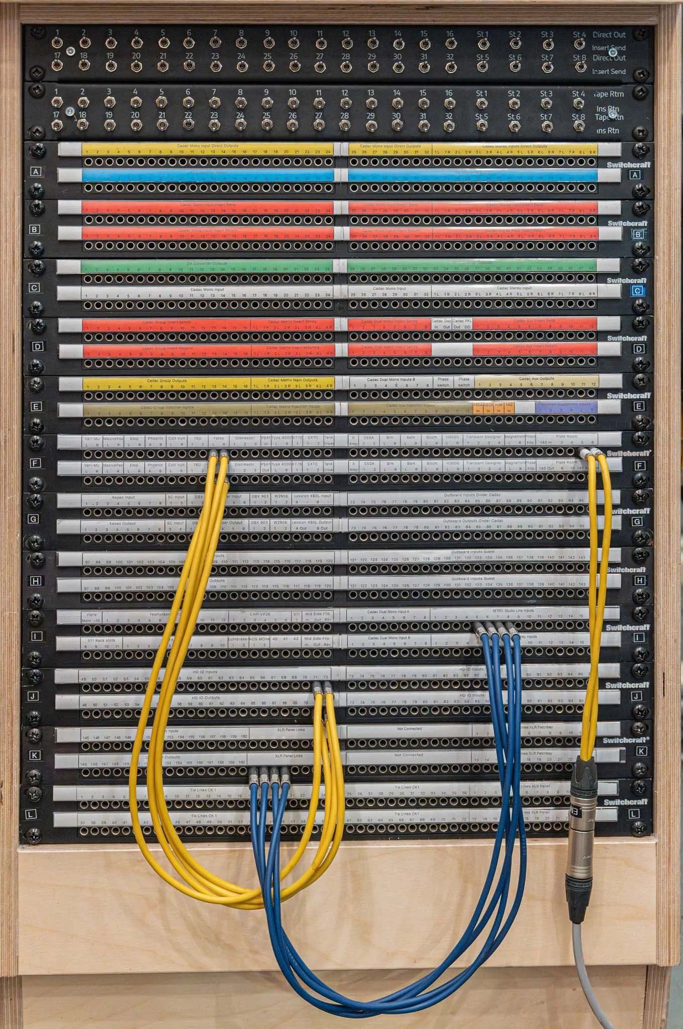 A large electronic audio mixing console with numerous color-coded input and output channels, sliders, and switches connected by yellow and blue cables.