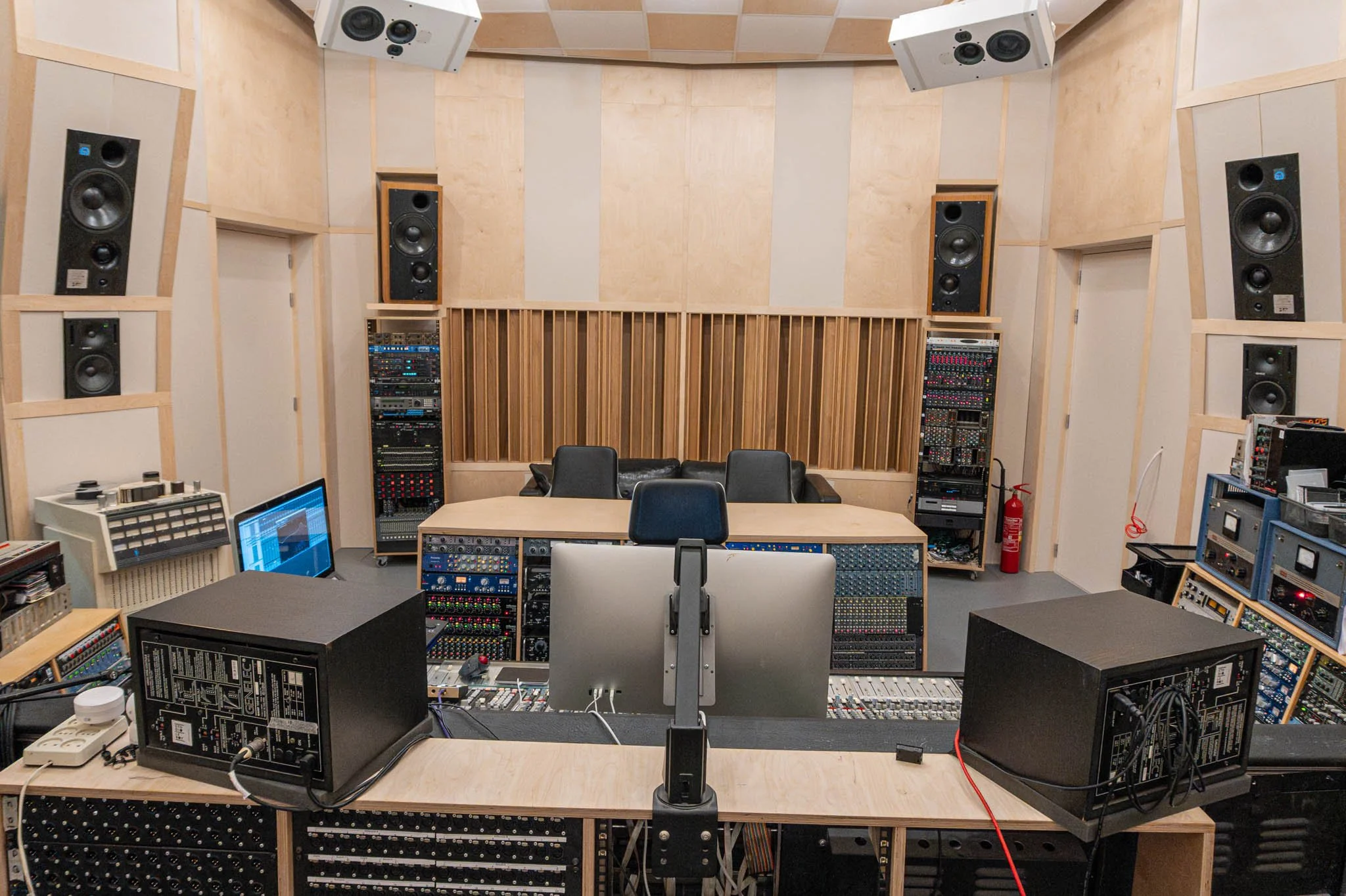 A professional recording studio with wooden soundproofing walls, multiple large speakers mounted on the walls, and various audio equipment and racks arranged around the room. In the center, there is a mixing console, computer monitor, and chairs.