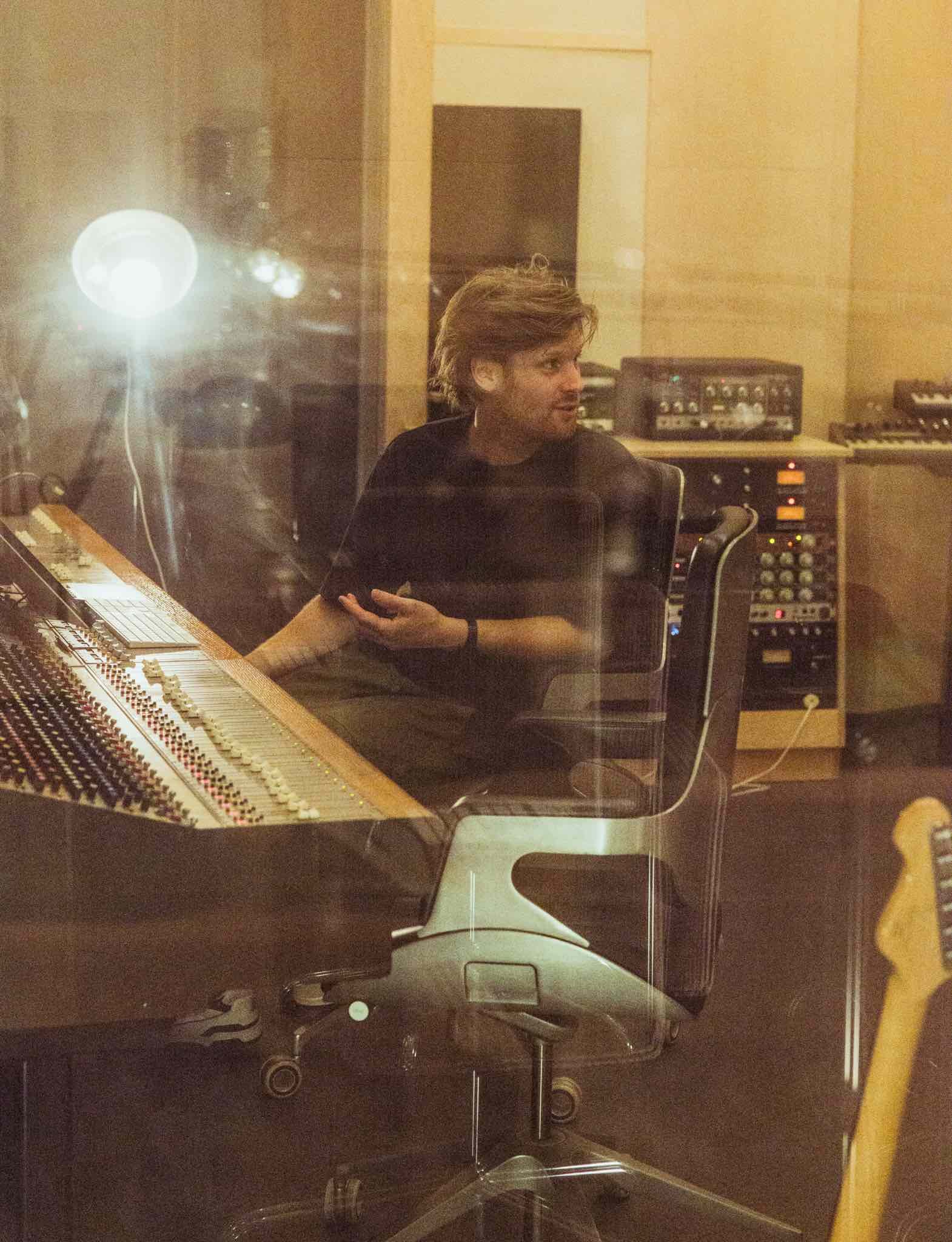 Person sitting in a recording studio, reflected through a glass window, with musical equipment and instruments in the background.