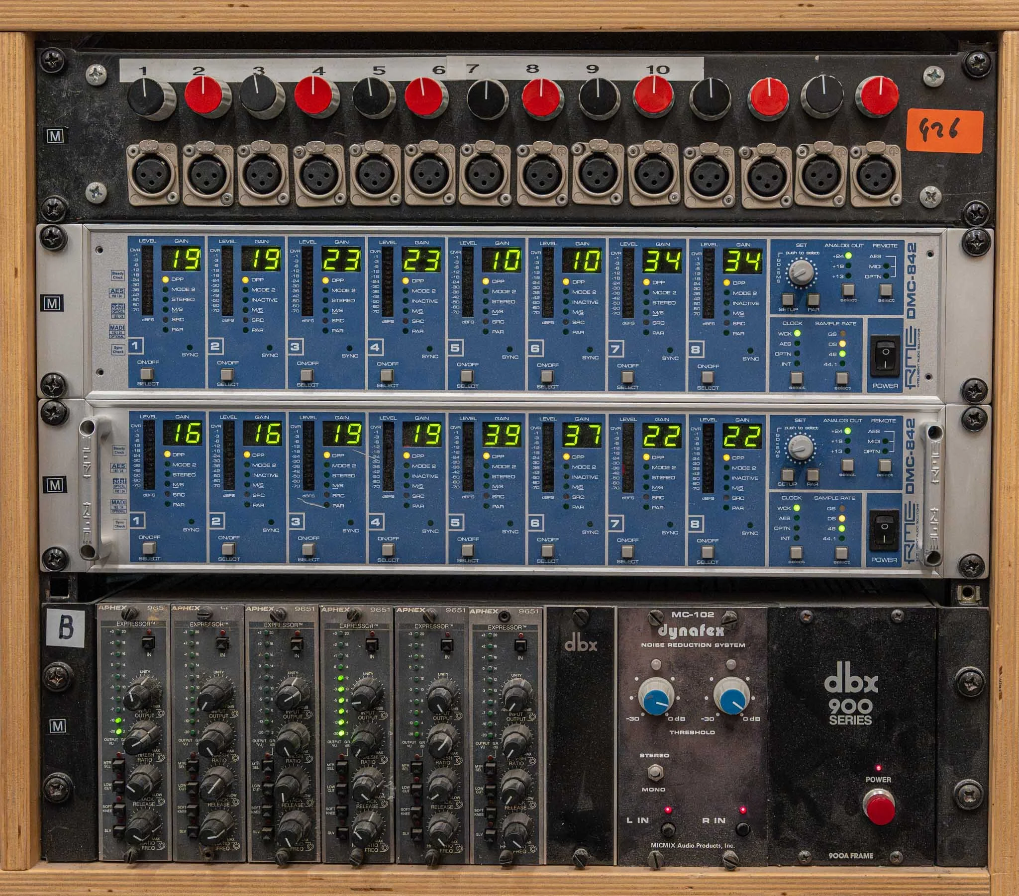 A professional audio rack with various audio equipment including equalizers, audio interfaces, and a noise reduction system, with colorful knobs and digital displays.