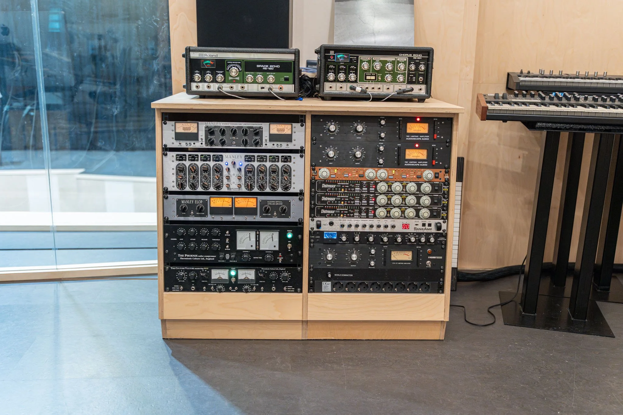 Audio recording studio equipment rack with various audio hardware units, including compressors, equalizers, and preamps, next to a keyboard on a stand.