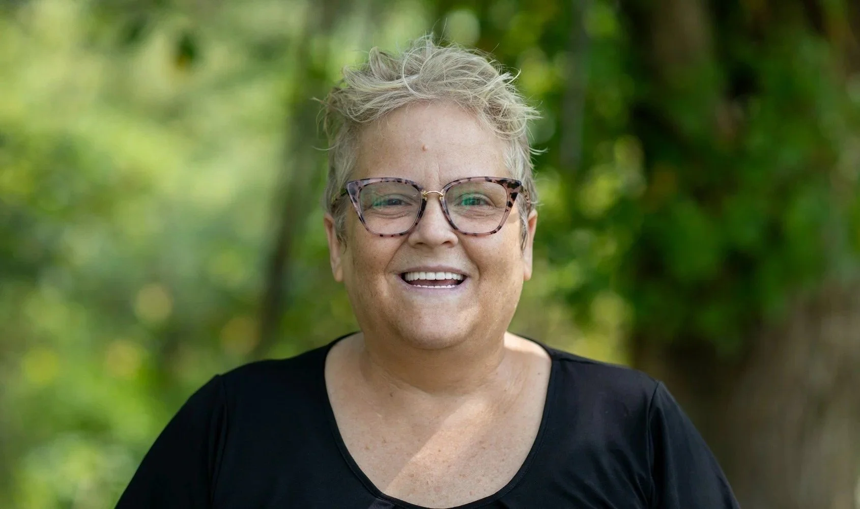 A smiling woman with short blonde hair, wearing glasses and a black top, outdoors with green blurred trees in the background.