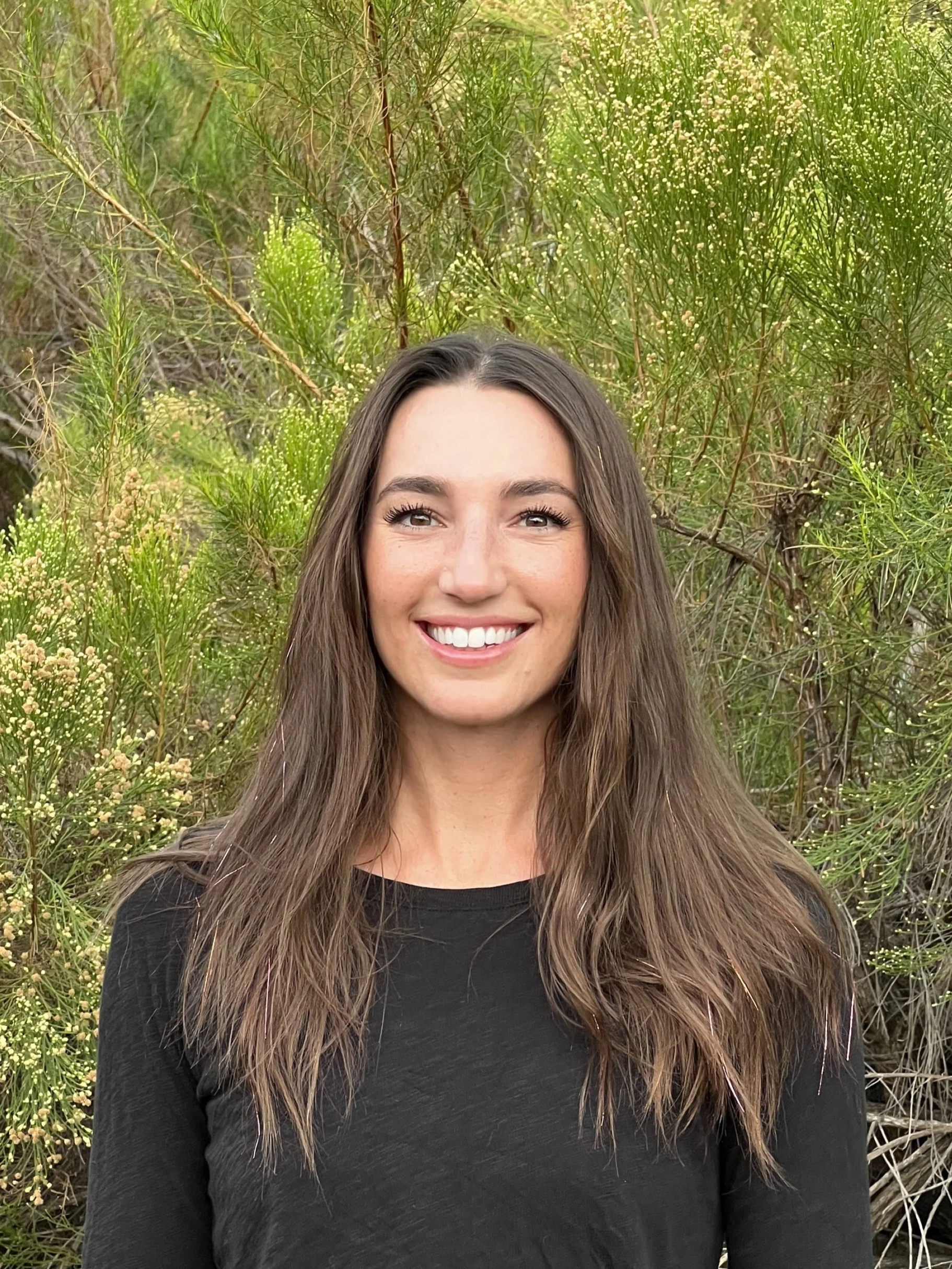 A woman with long brown hair smiling in front of green shrubbery and small white flowers. The woman is the owner/funder. The shrub is Baccharis, an important food source for pollinators and birds in the late summertime when food is scarce.