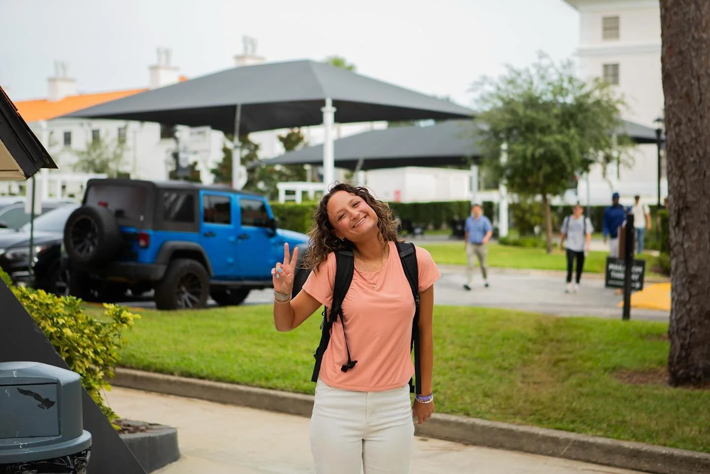 A smiling young woman with curly hair making a peace sign with her right hand, standing outside in a parking lot with green grass and trees, wearing a peach-colored shirt, white pants, and a backpack, with several people in the background.