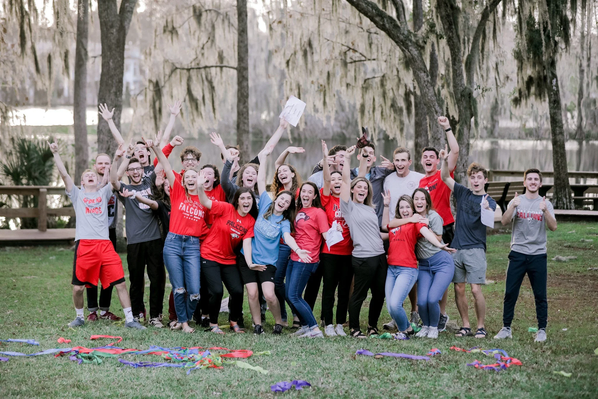Group of cheerful young people outdoors celebrating, some wearing red and gray shirts, with trees and a lake in the background.