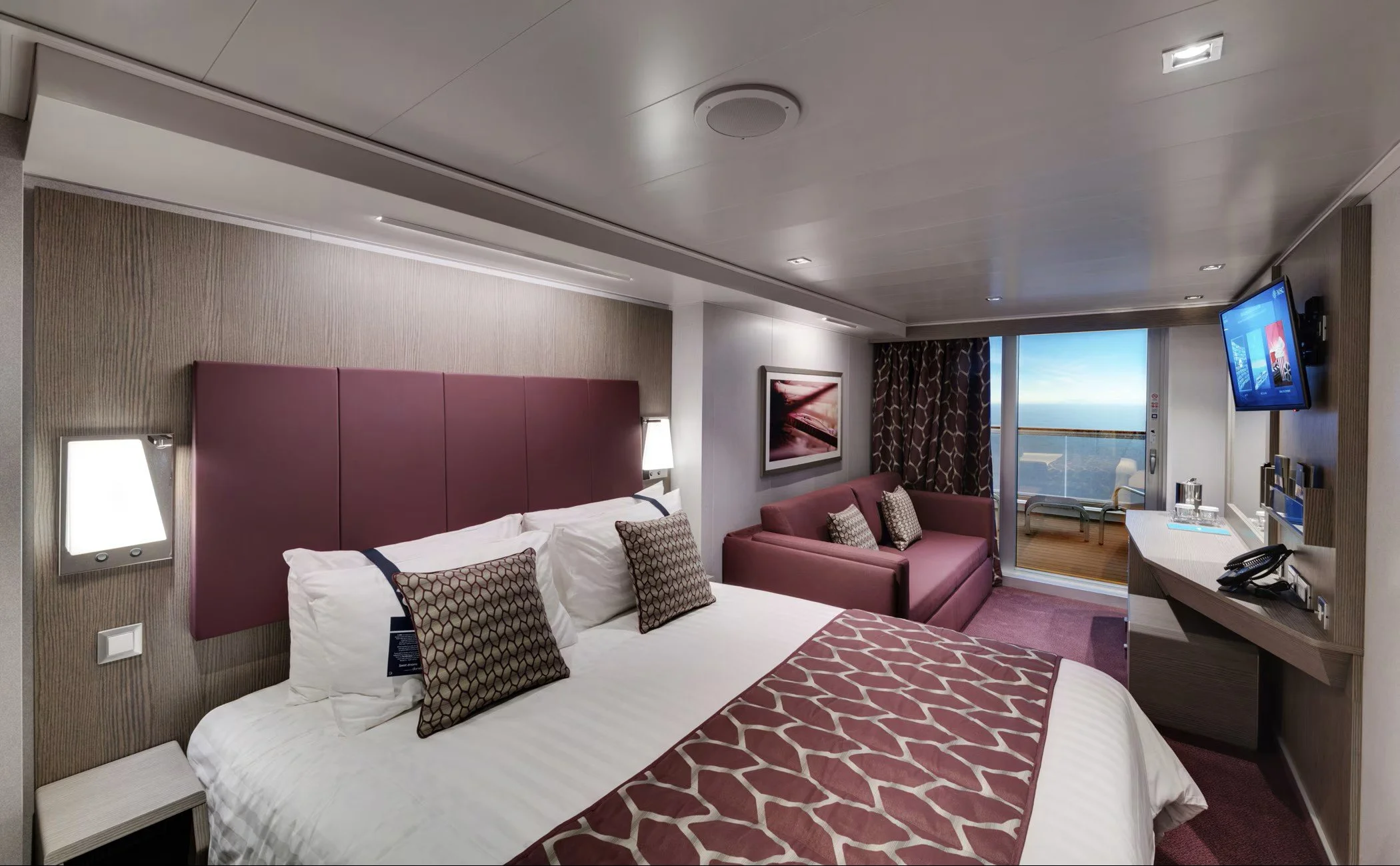 A cruise ship hotel room with a bed, sofa, wall-mounted TV, balcony door, and ocean view.