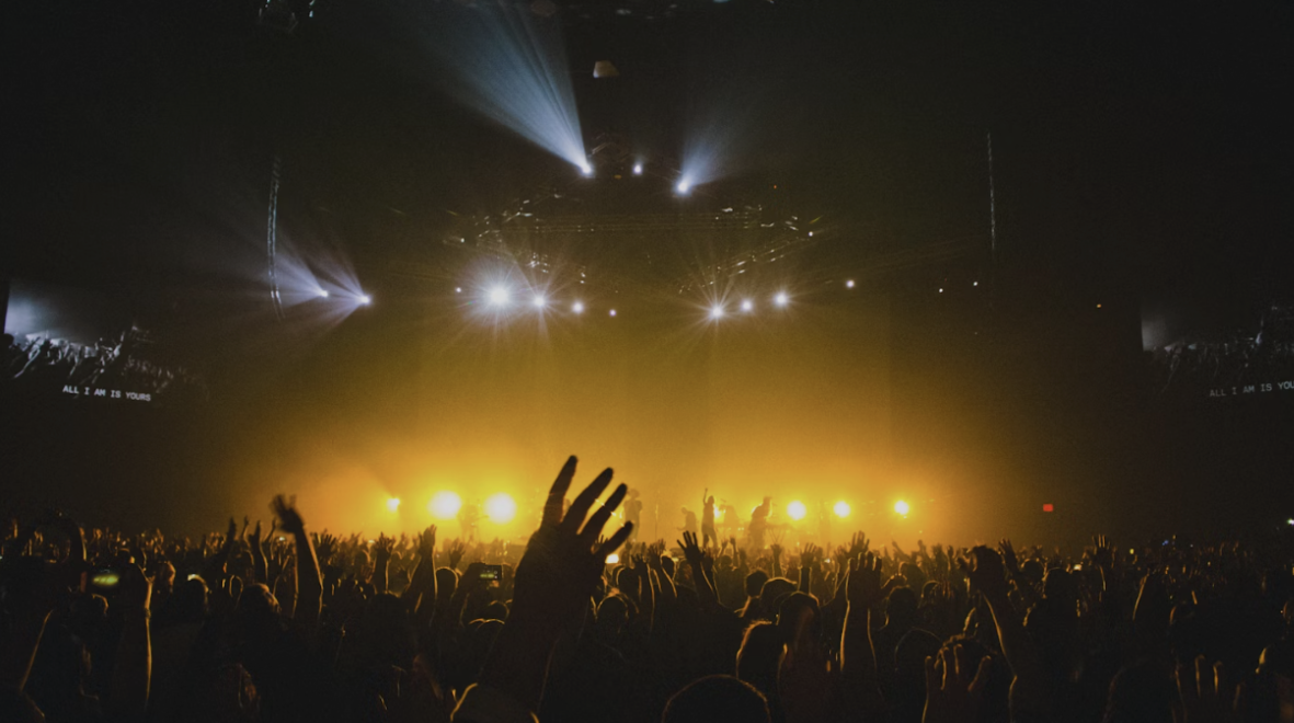 Crowd at a concert with bright stage lights and raised hands.