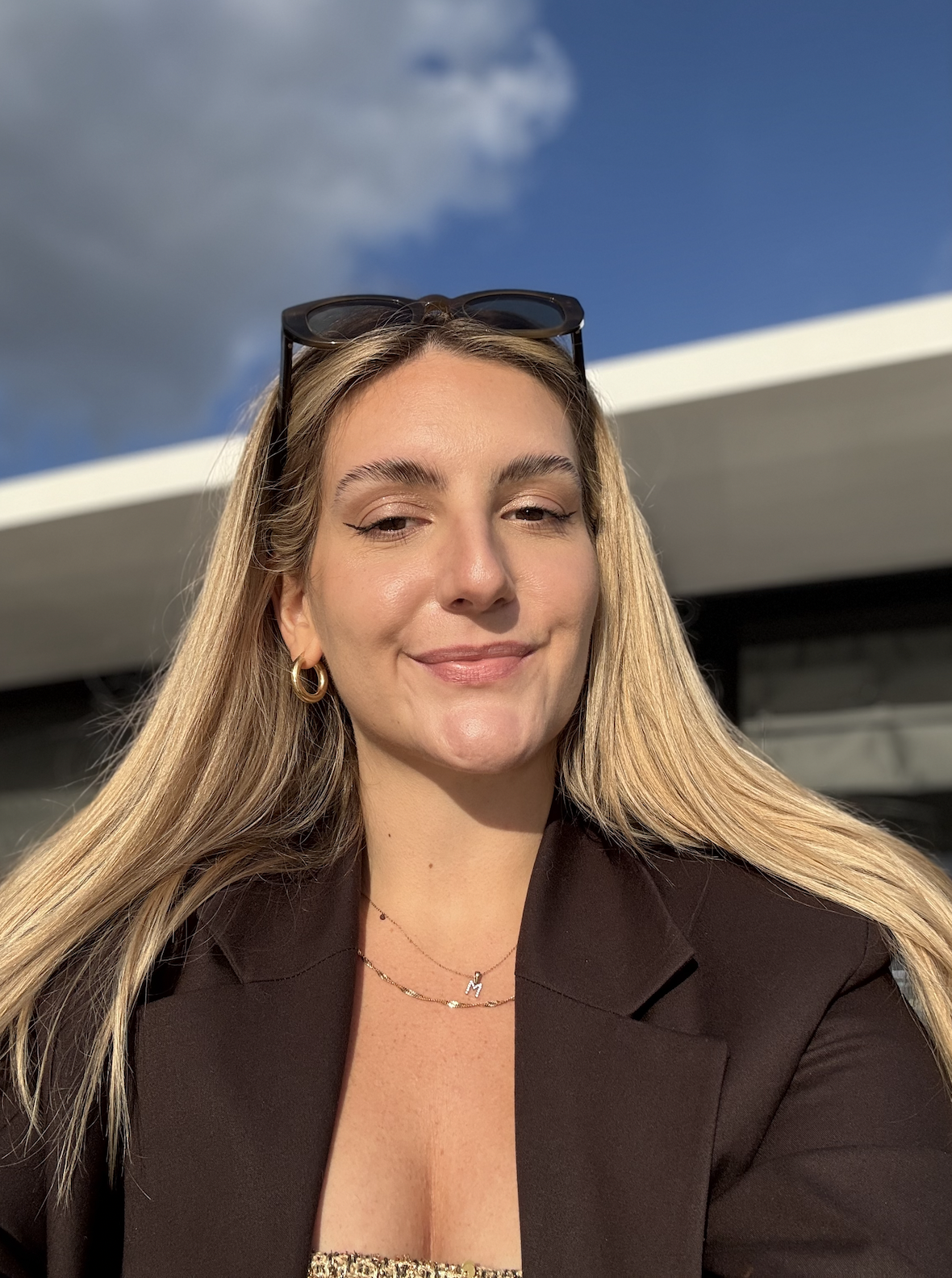 Monika Raszowska. A woman with long blonde hair outdoors, wearing black sunglasses on her head, gold hoop earrings, a black blazer, and layered necklaces, smiling with a partly cloudy sky in the background.