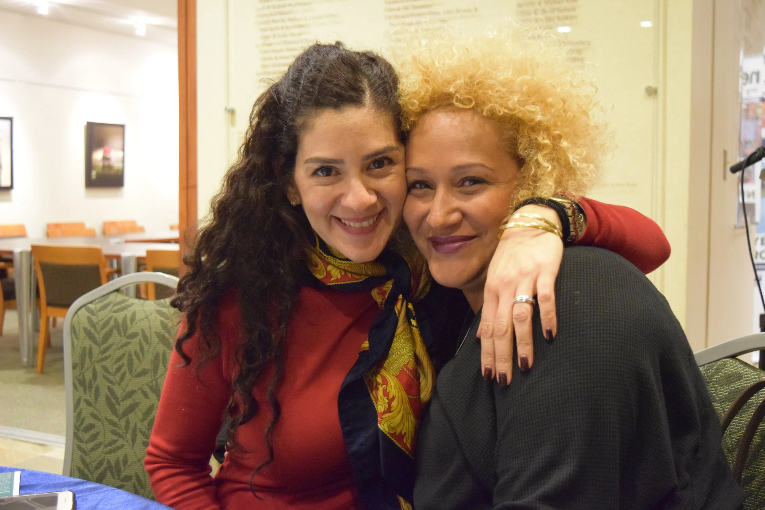 Two women hugging and smiling at the camera in an indoor setting, with chairs and artwork on the walls in the background.
