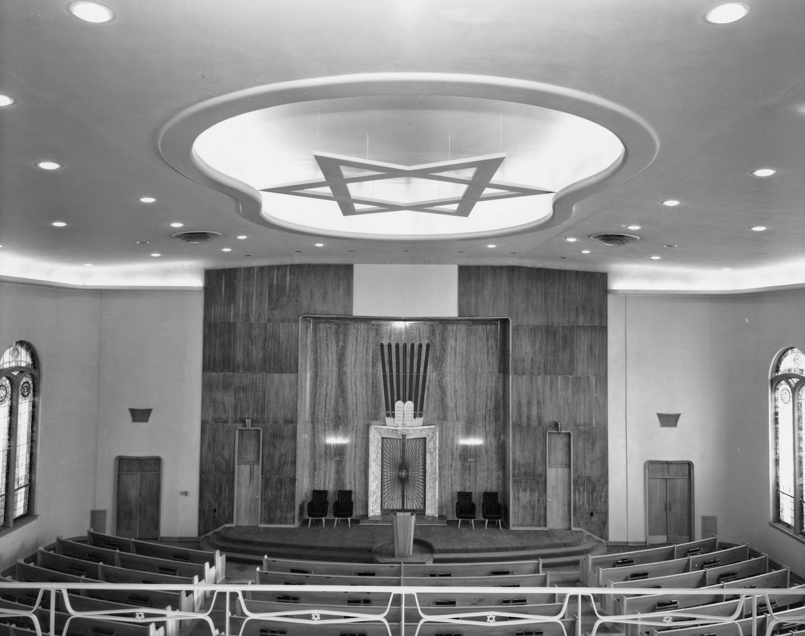 Inside view of a church or synagogue with an altar at the front, wooden pews, stained glass windows, and a Star of David symbol on the ceiling.