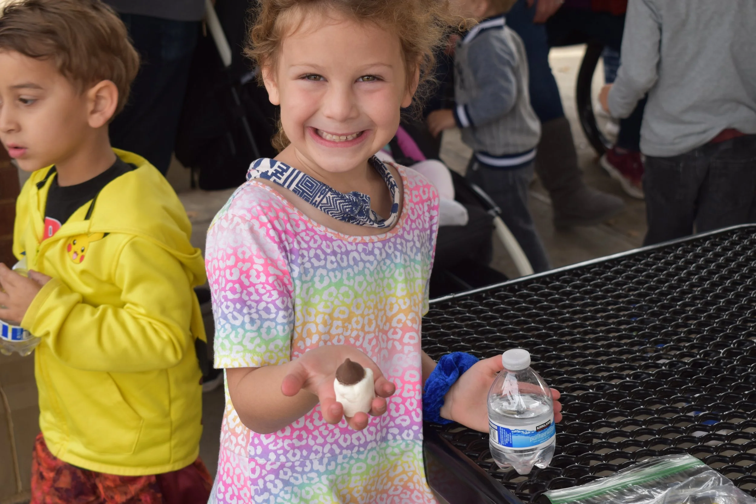 A young girl with curly red hair smiling and holding a marshmallow and a water bottle, surrounded by other children at a social gathering.