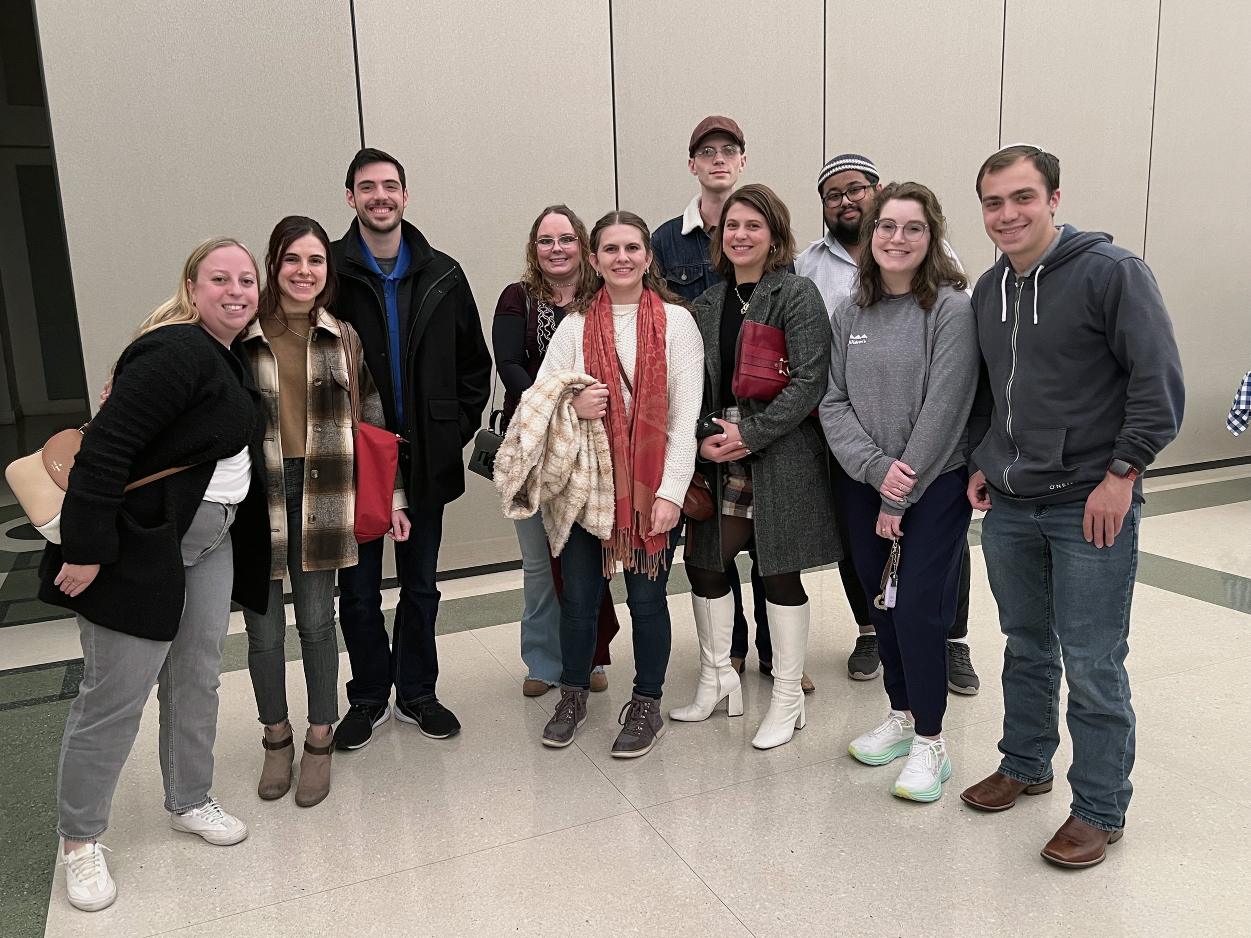 Group of eleven people posing for a photo indoors against plain beige walls, dressed in casual and business casual clothing, smiling and standing close together.