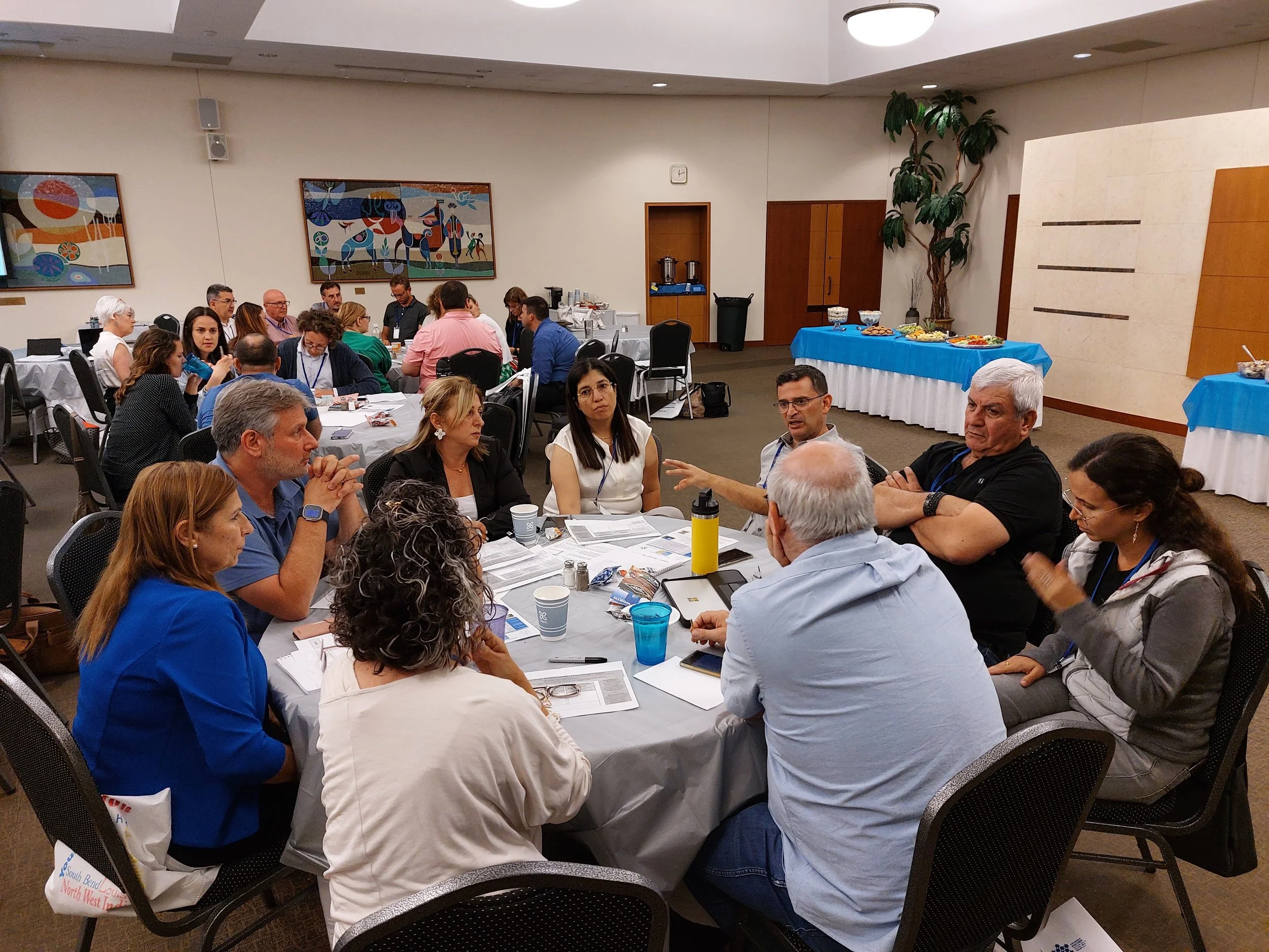 People in a conference room engaged in a discussion around a table, with a buffet table at the side and artwork on the walls.