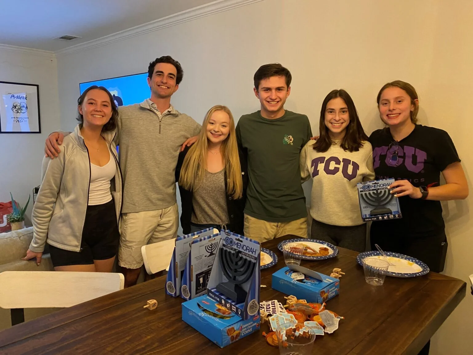 Seven young adults standing at a dining table smiling at the camera, with holiday treats and a menorah-themed game on the table.