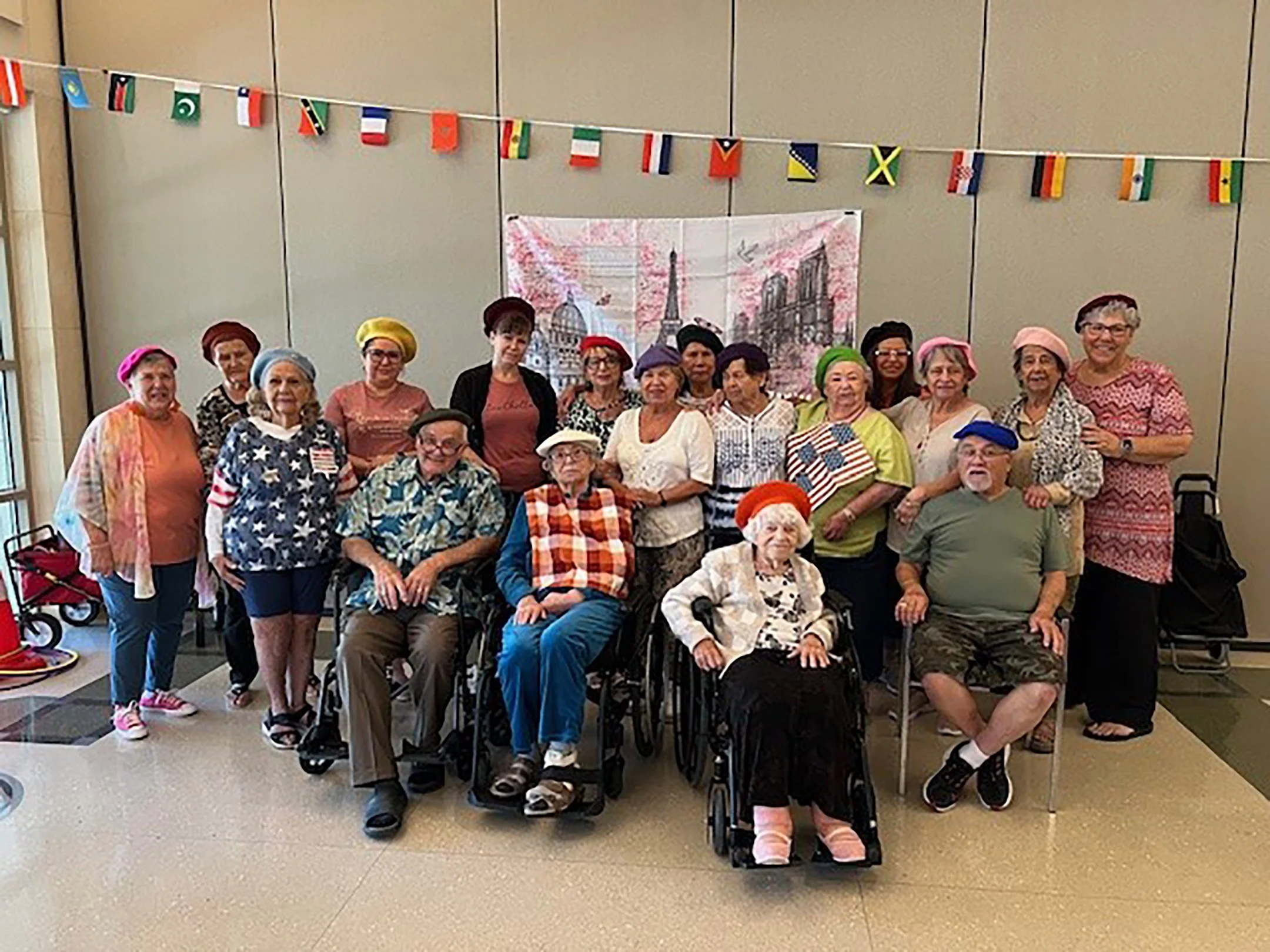 Group of seniors, some in wheelchairs, in patriotic clothing and hats, gathered indoors for a celebration with American flags and a backdrop of Paris.