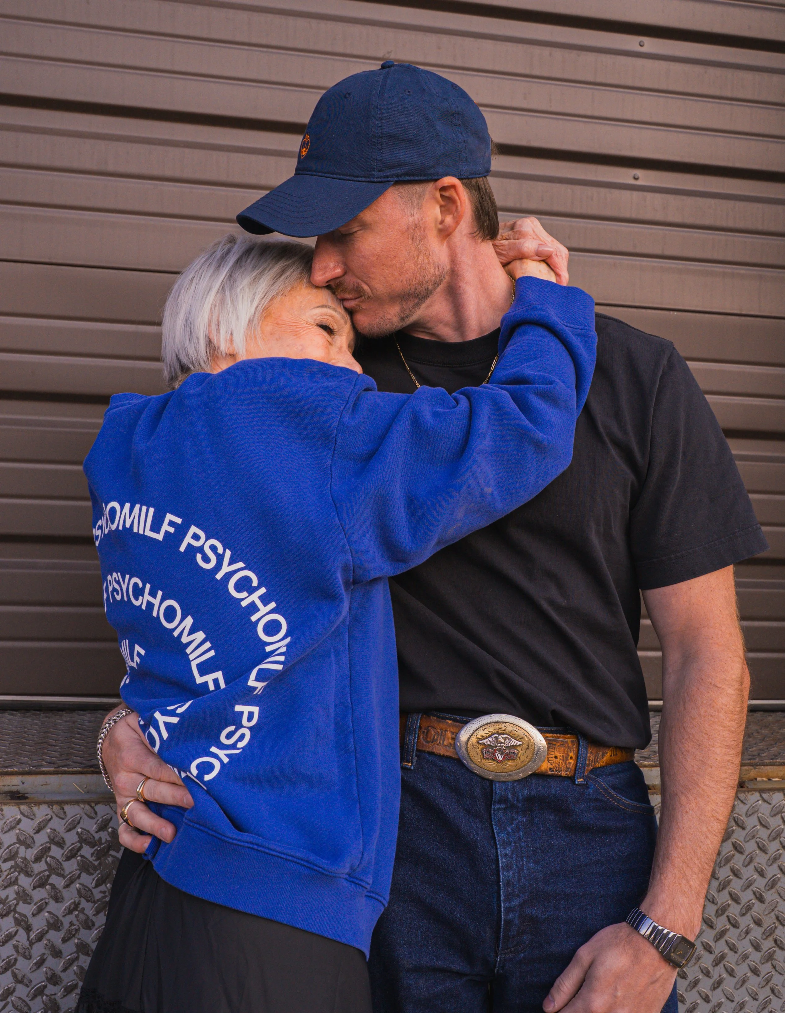Older woman and younger man embracing, the man is wearing a blue psychomilf embroidered baseball hat and the woman is wearing a blue psychomilf crewneck