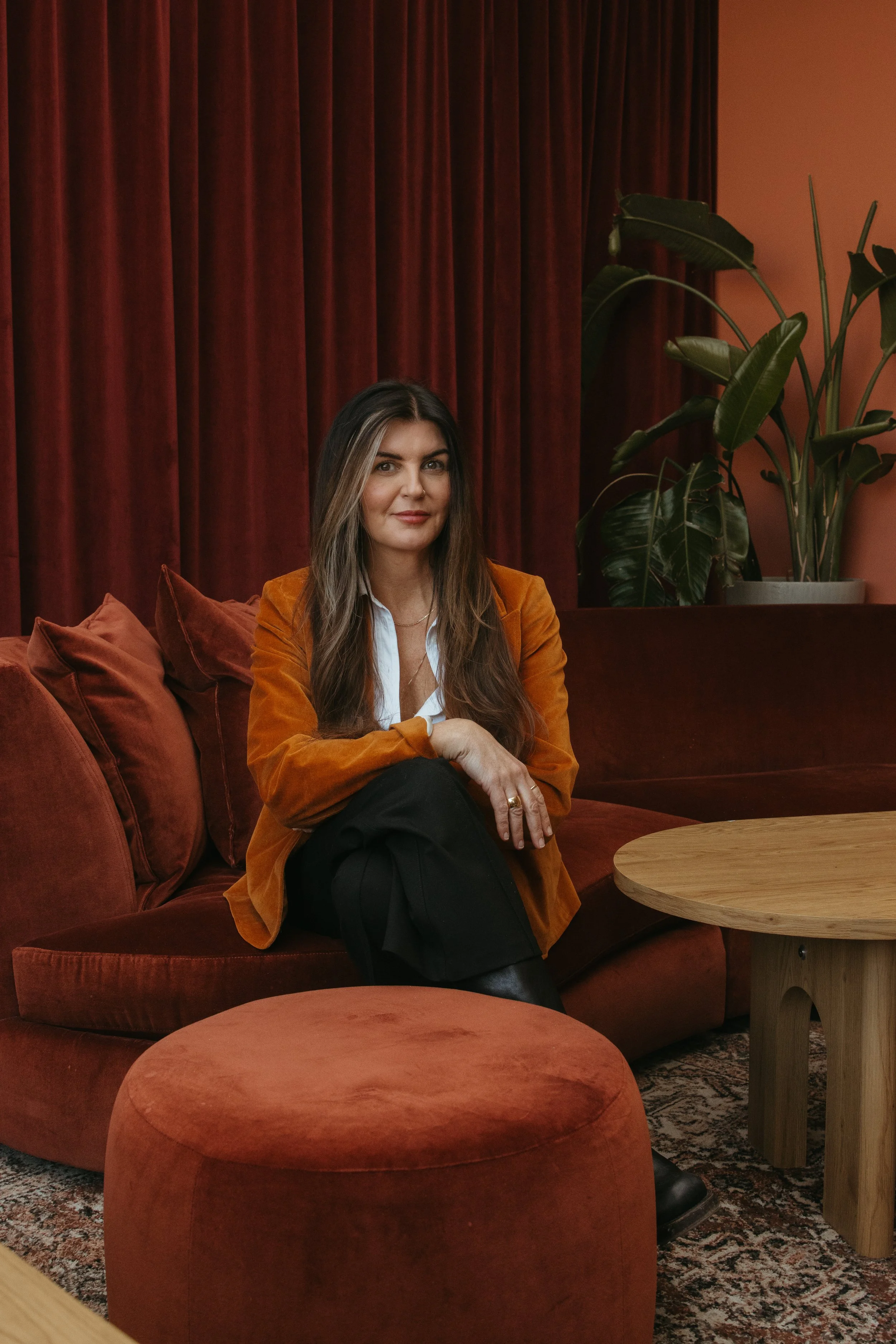 A woman with long brown hair and a white shirt sitting on an orange velvet sofa in a room with red curtains and a large green plant.