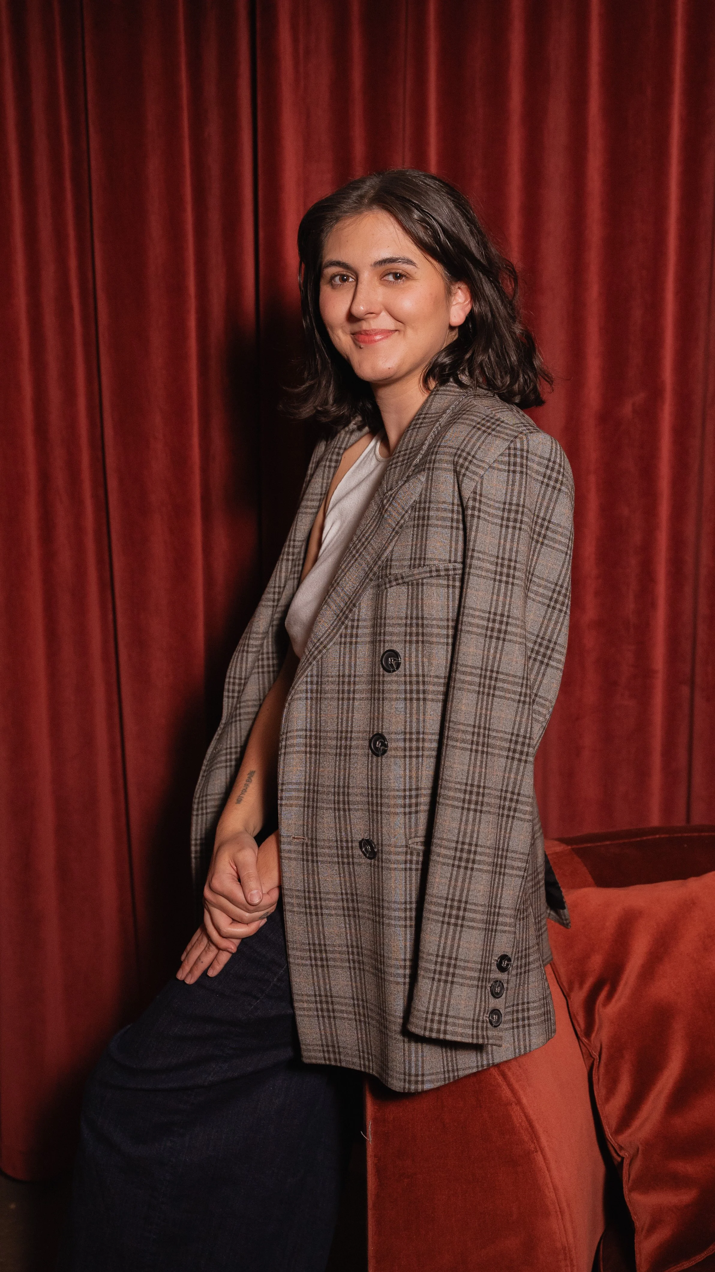 A woman with shoulder-length dark hair and a light smile, standing in front of a red curtain, wearing a checked blazer and casual clothes.