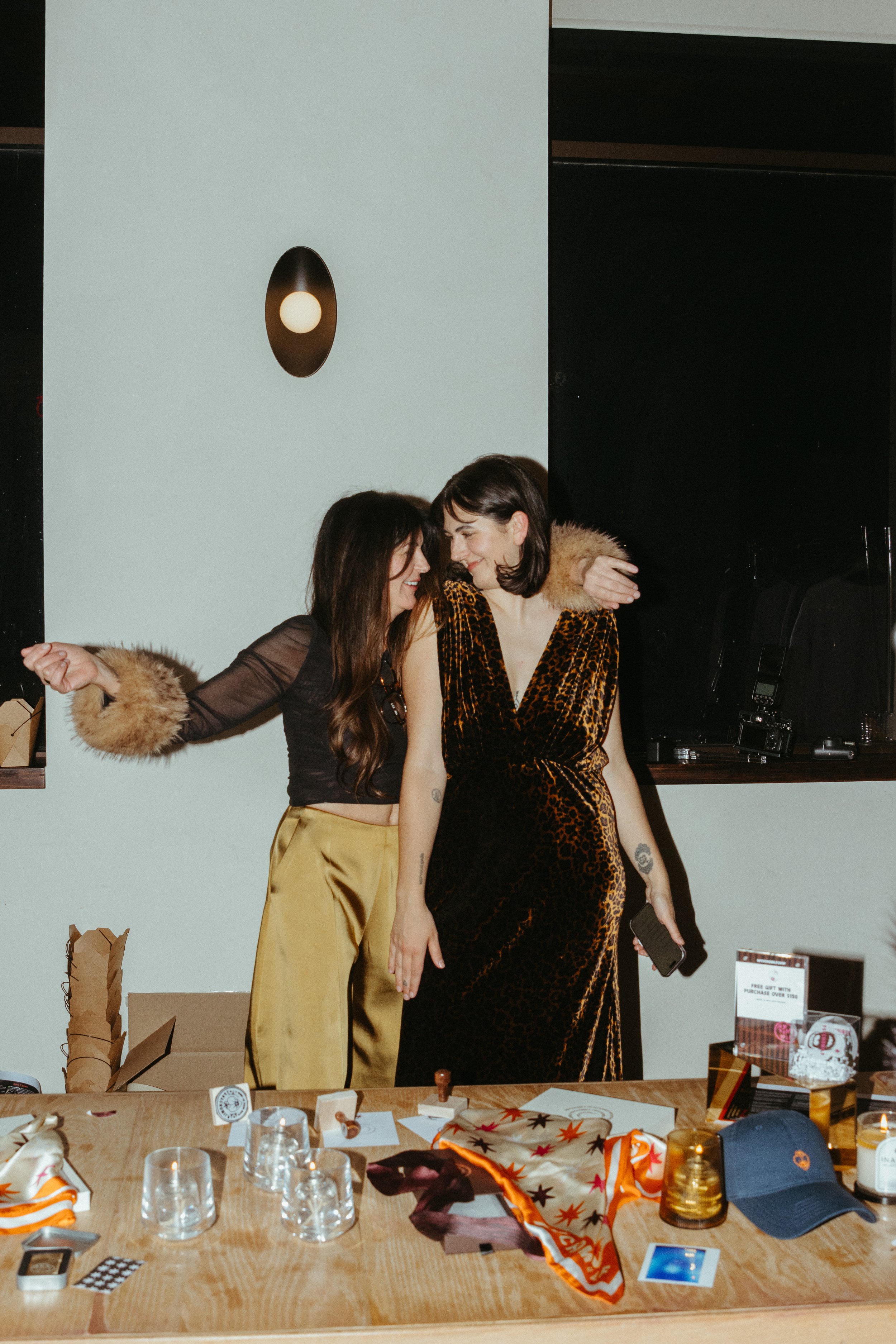 Two women smiling and embracing each other at a social gathering or party, standing behind a table with various items including glasses, a cap, and decorations.