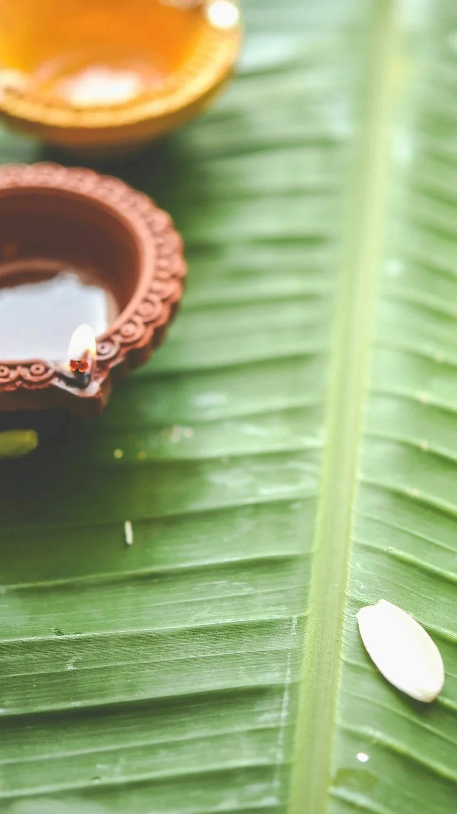 Close-up of green banana leaf with two traditional oil lamps made of clay, one with a small candle and the other filled with oil, and a white seed or petal on the leaf.