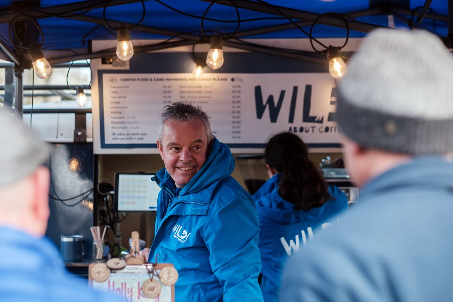 Founder, Phil Corin in a blue jacket serving coffee, talking to customers, with a menu and warm lighting overhead.