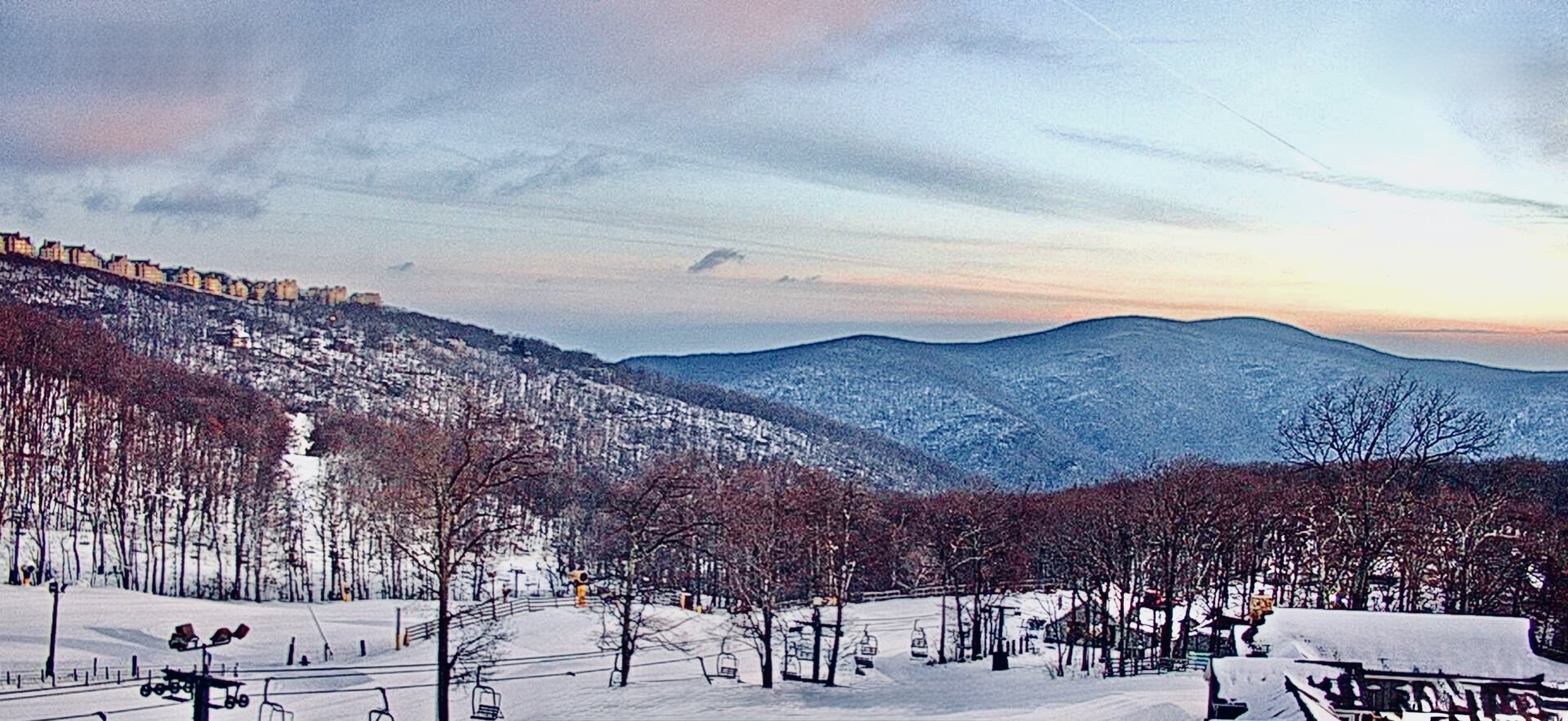 Snow-covered ski resort with ski lifts, trees, and mountain slopes during sunset or sunrise.